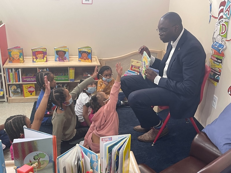 A man wearing a dark suit holds a book up to show young students sitting on a rug in a classroom. Some of the students are holding up their hands.