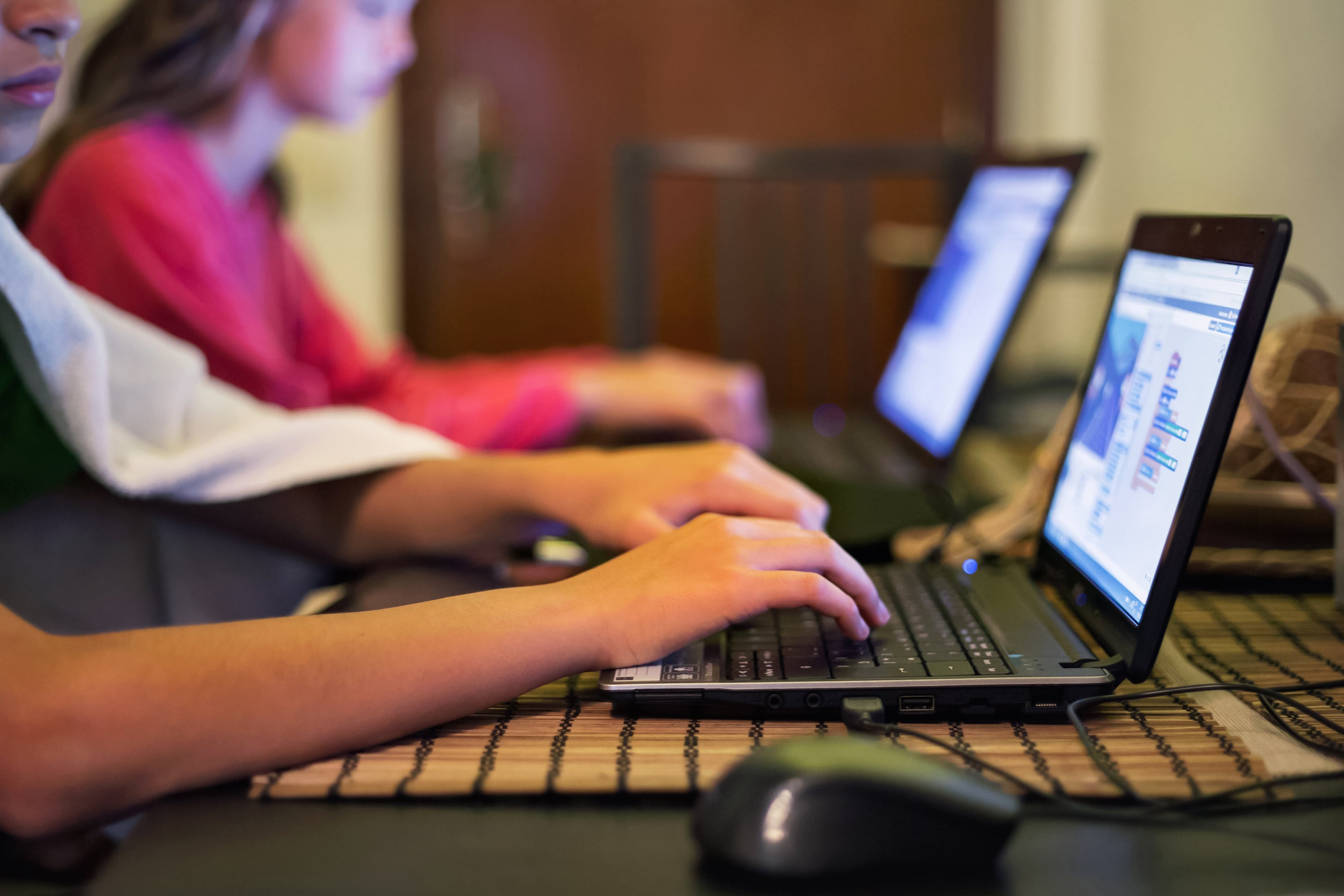 Two young students sit side by side and reach out to work on laptops. The student in the background is not in focus.