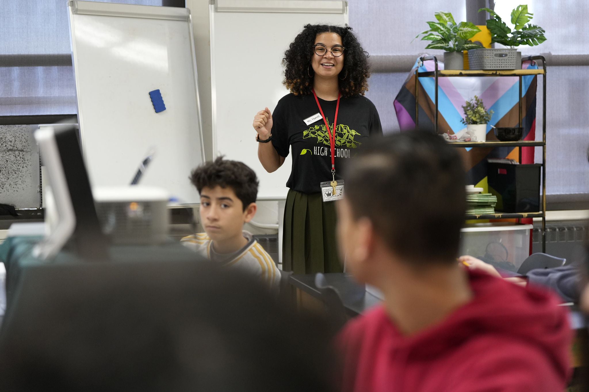 A teacher with medium length dark hair stands next to two students in a classroom with dry erase boards and flags in the background.