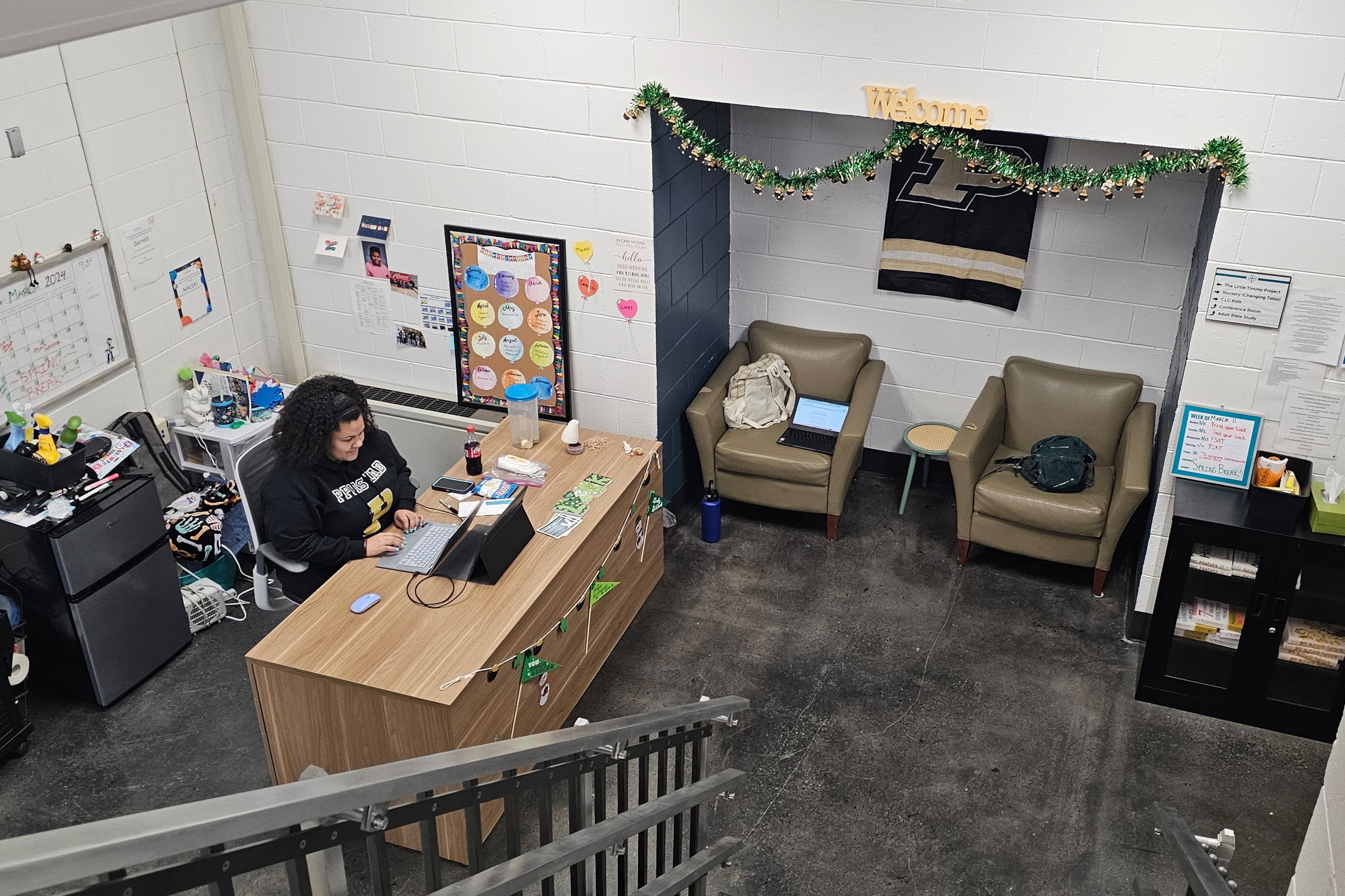 A view from above of an adult with dark hair and wearing a dark sweater sits at a wooden desk typing at a laptop in an office environment.