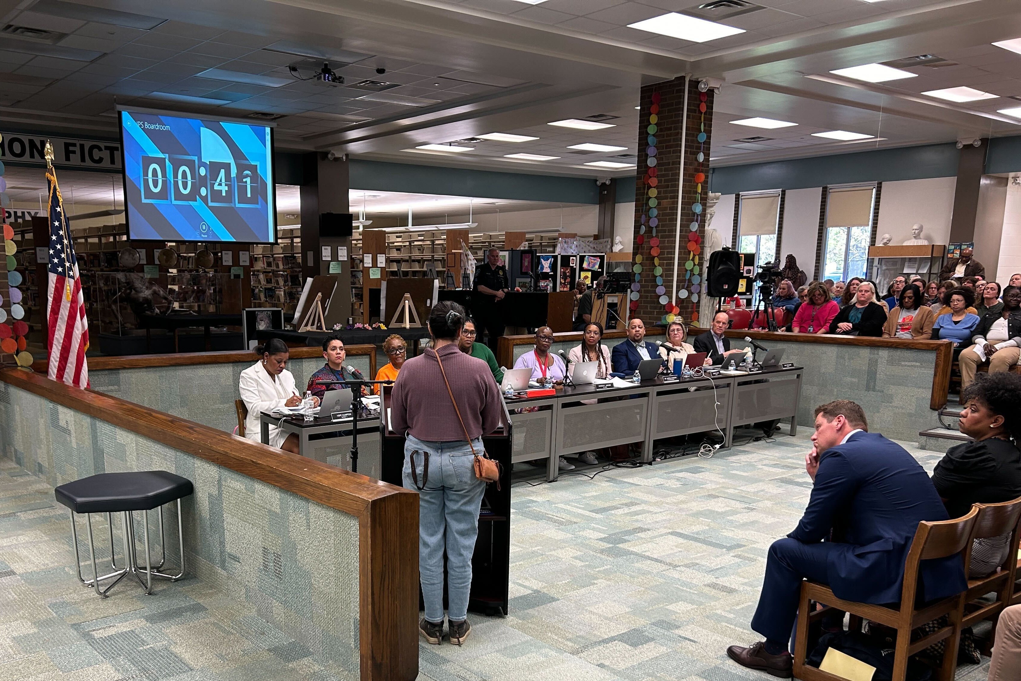 A person stands at a podium in front of a row of people sitting at a long desk with people sitting in the foreground.