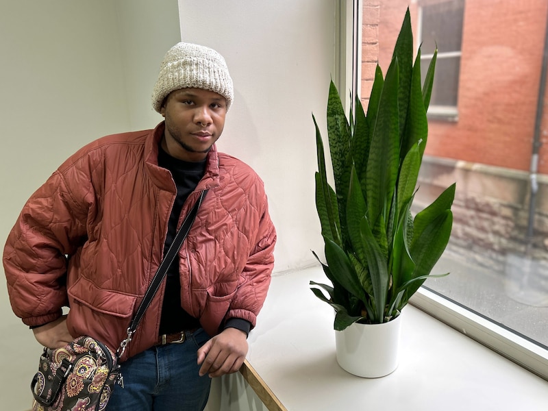 A young man poses for a portrait by a window and a green plant while wearing a white hat and a red jacket.