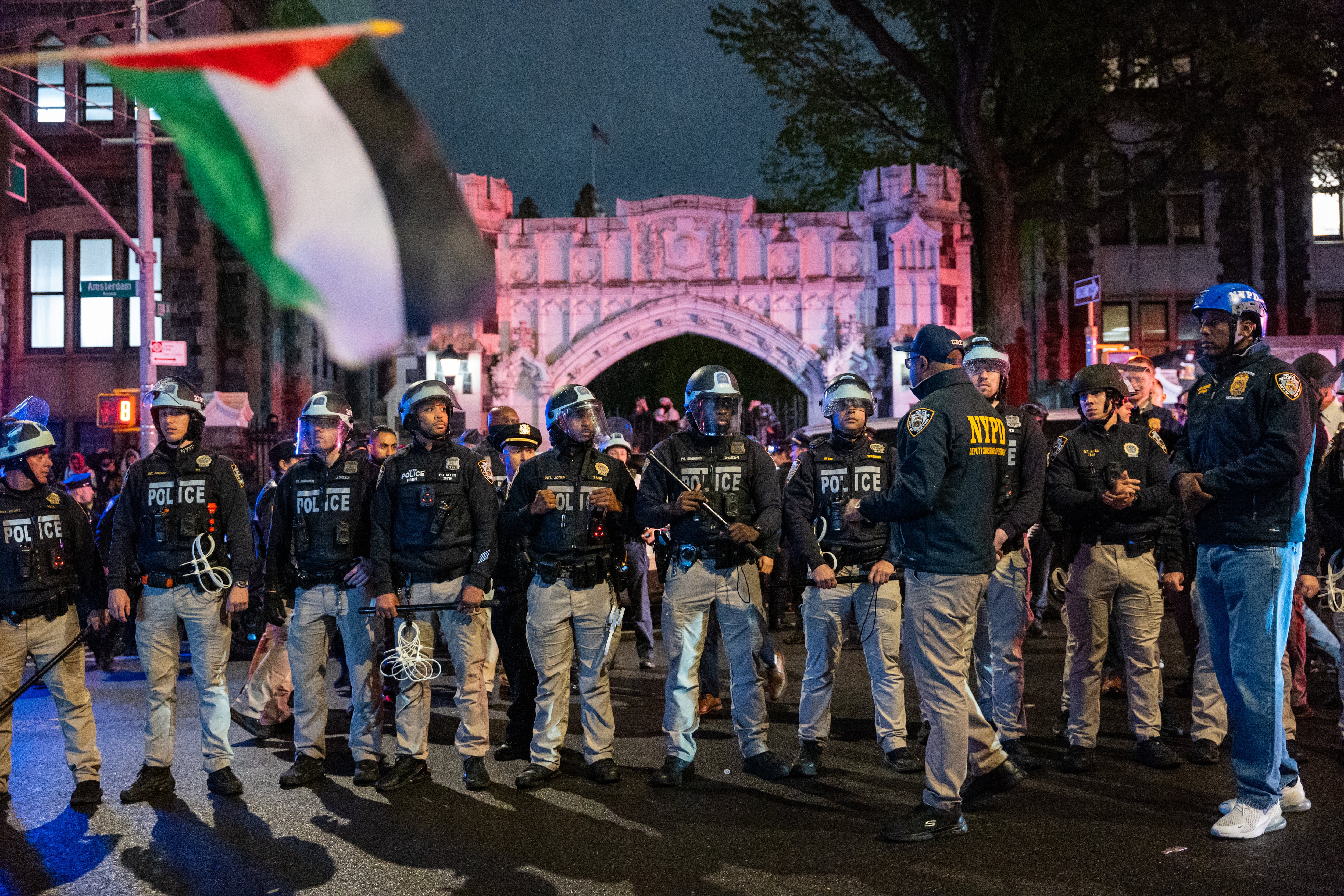 A line of police officers stand in front of an arch with a Palistinian flag in the foreground.