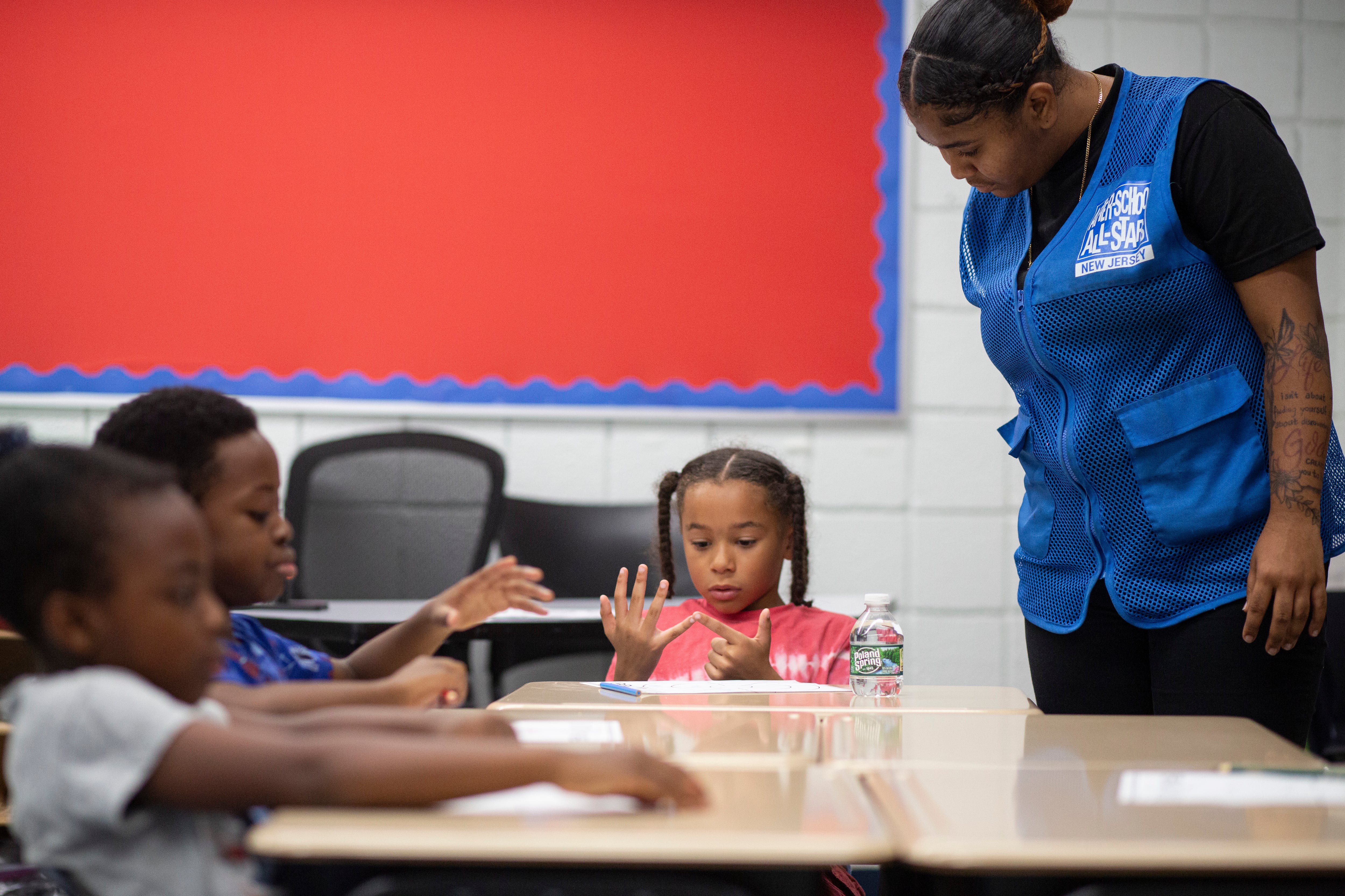 A adult stands above a desk full of young students working on math.