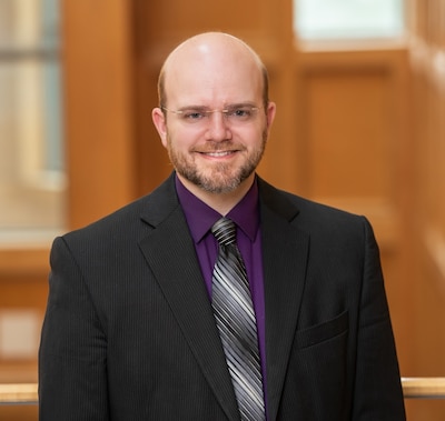 Headshot of a man wearing a purple shirt, a black blazer, and a black and white tie.