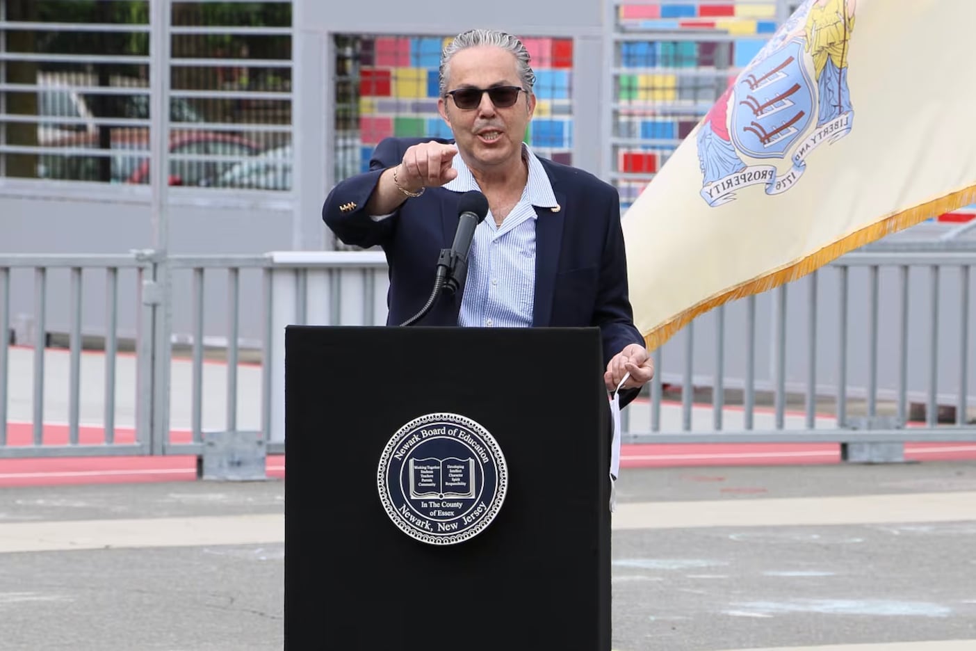 A man wearing sunglasses and a suit jacket points forward while standing at a podium with a colorful building and a flag in the background.