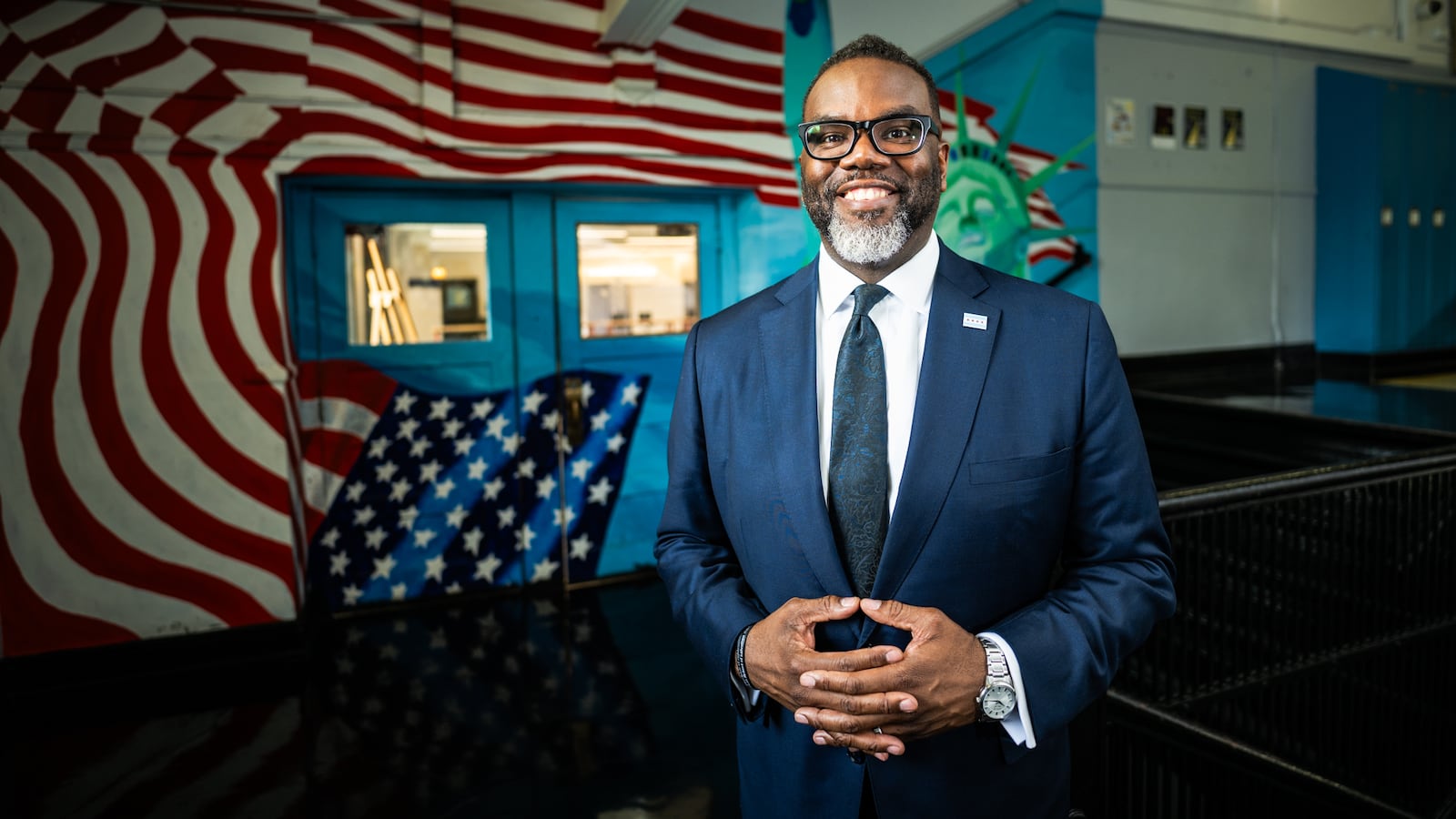 Chicago Mayor Brandon Johnson smiles while standing in a school hallway with a mural of an American flag behind him.