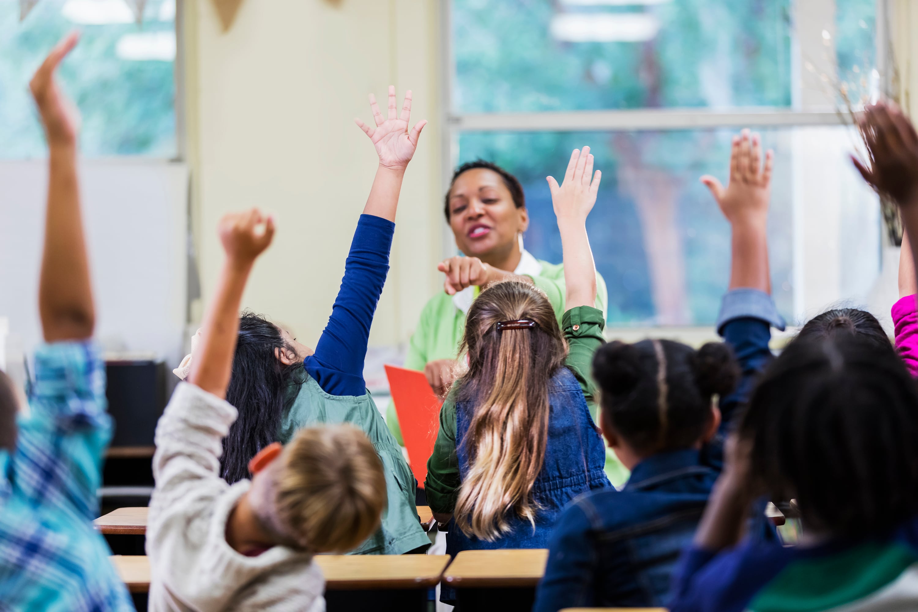 A view of a classroom from the back of the room with students raising their hands and a teacher in the background calling on someone.