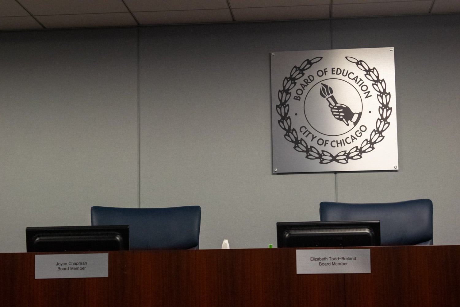 A photo of two empty seats for Chicago’s school board members on shared dais. In front of the seats is the table part of the dais. Behind the seats is a gray, matte, and square sign. In the middle of the sign is a circle with a hand holding a torch inside. Above the circle is the words “Board of Education,” curved around the circle. Below is the circle is the words “City of Chicago,” also curved. Wrapping around the words in a half circle is a laurel.