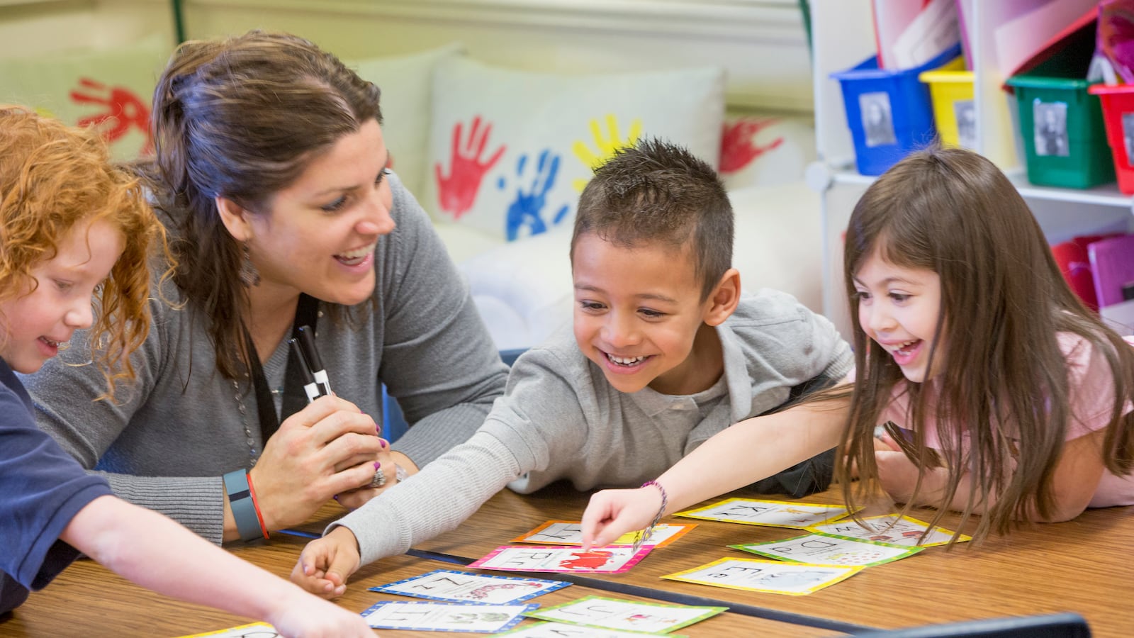 Young students surround their teacher at a classroom table. They are learning their letter.s