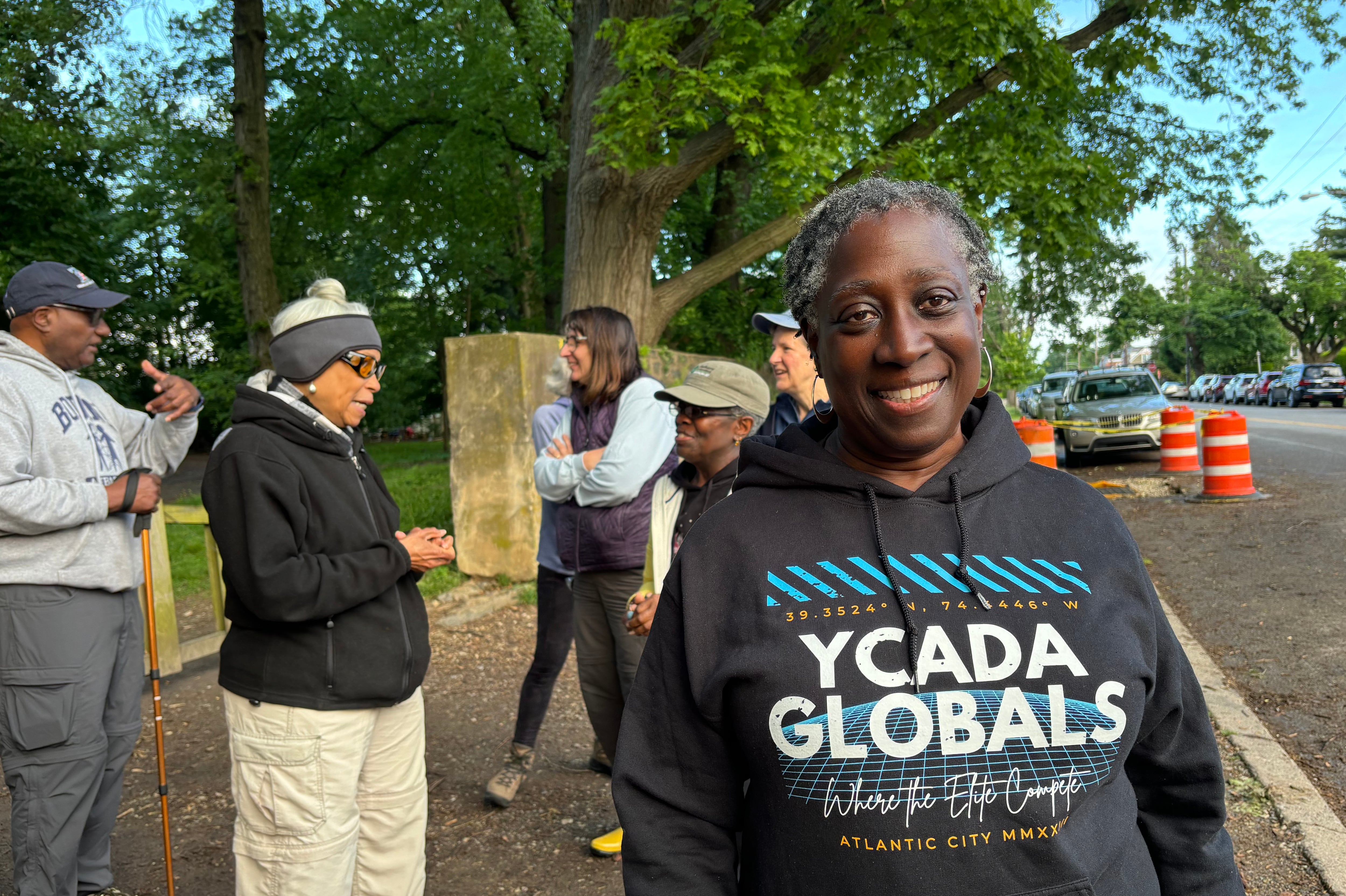 A woman with short hair and wearing a black sweater with writing on it stands outside for a portrait with other people in the background.