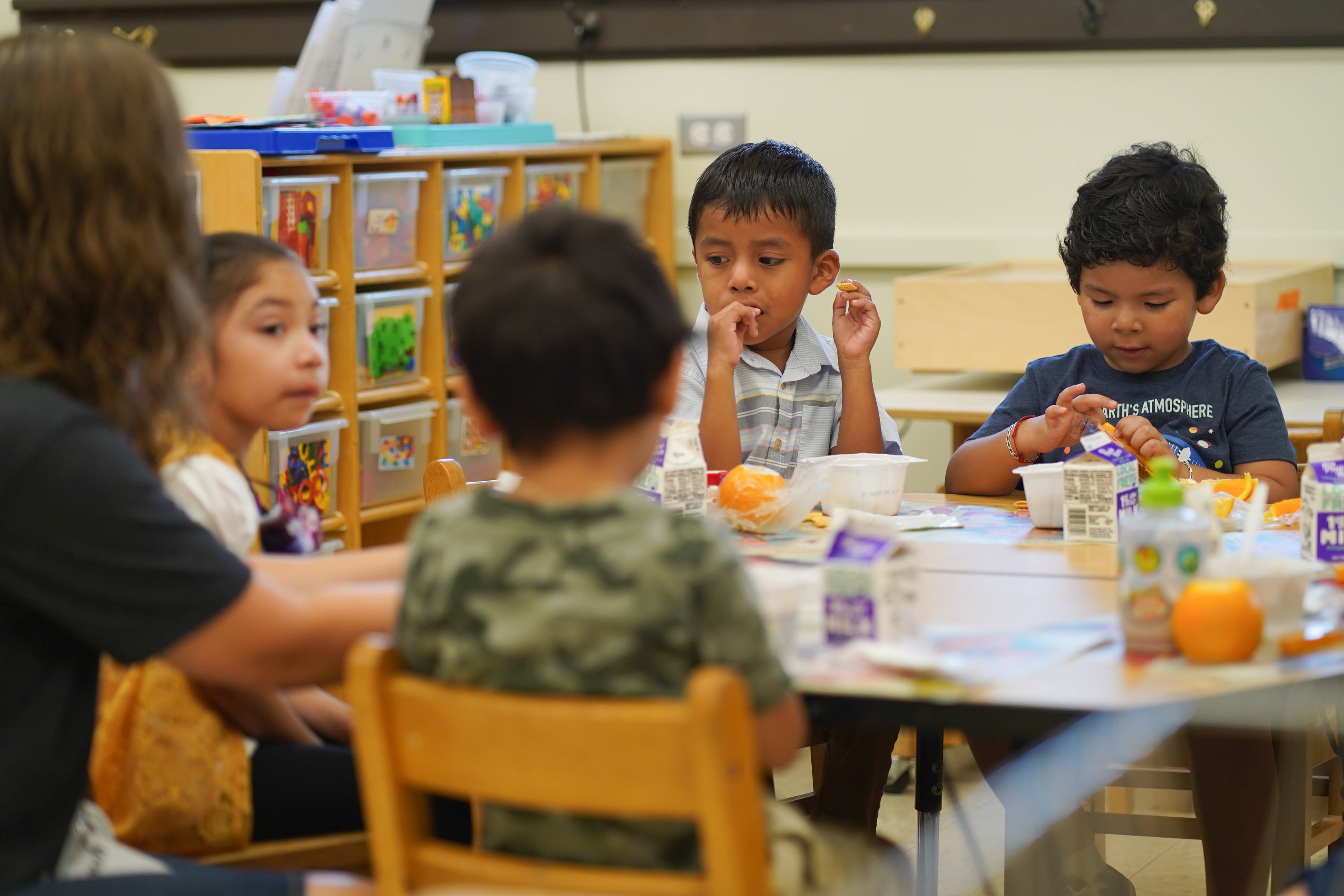 Four young students and an adult sit at a table in a classroom. There are toys and a white wall in the background.