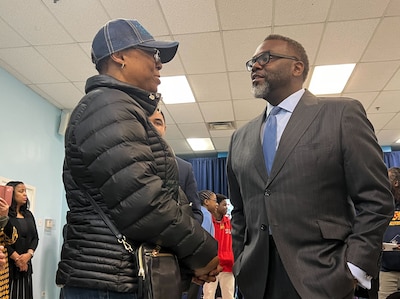 Two adults, one wearing a black winter coat and one wearing a black suit jacket, talk with each other with other people in the background.