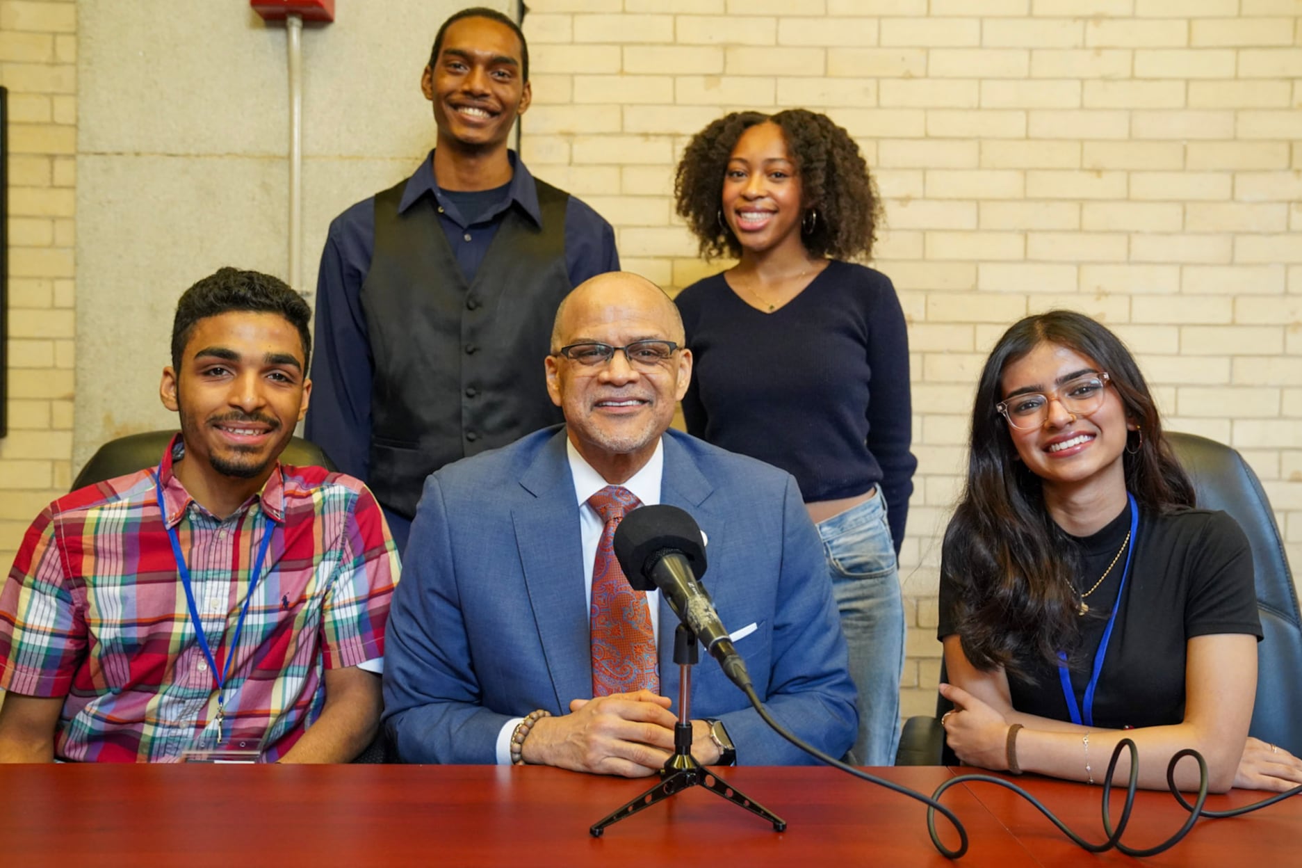 An adult man in a blue suit sits in the middle of student high school students at a wooden desk while two other high school students stand in the back all posing for a portrait.