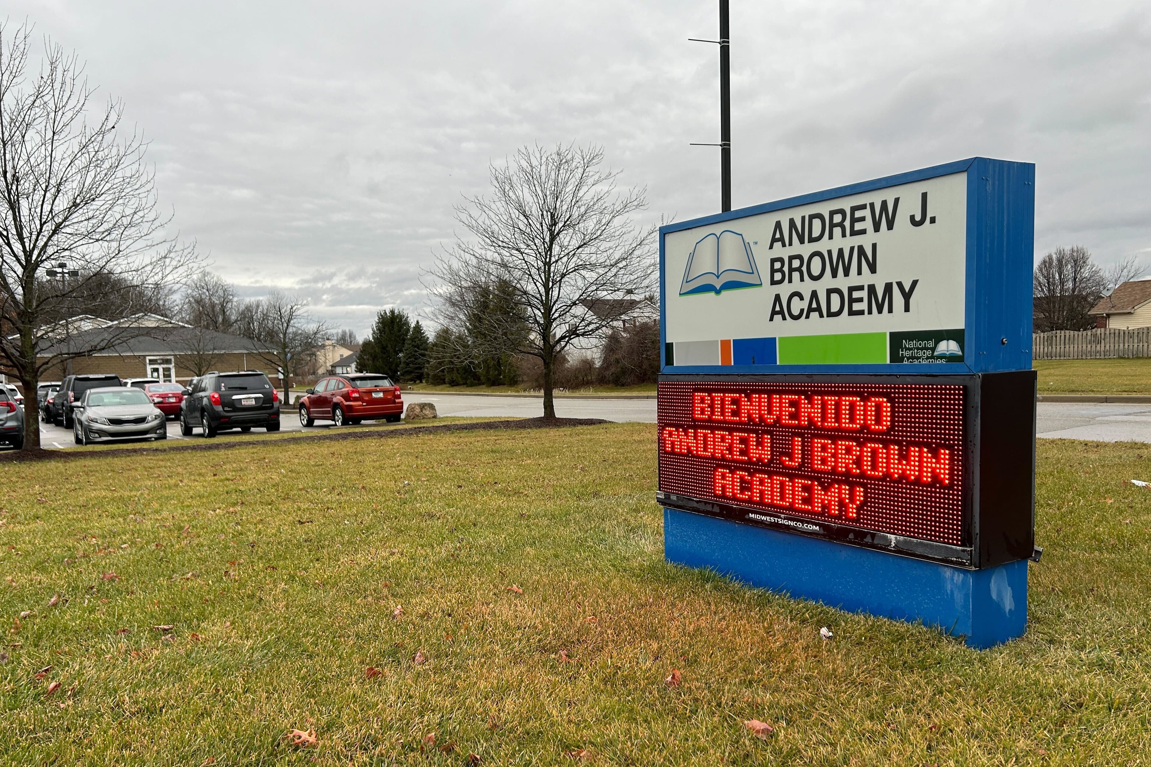 A sign that reads "Andrew J. Brown Academy" and "Bienvenidos Andrew J. Brown Academy," sits on a green lawn with cars and grey sky in the background.