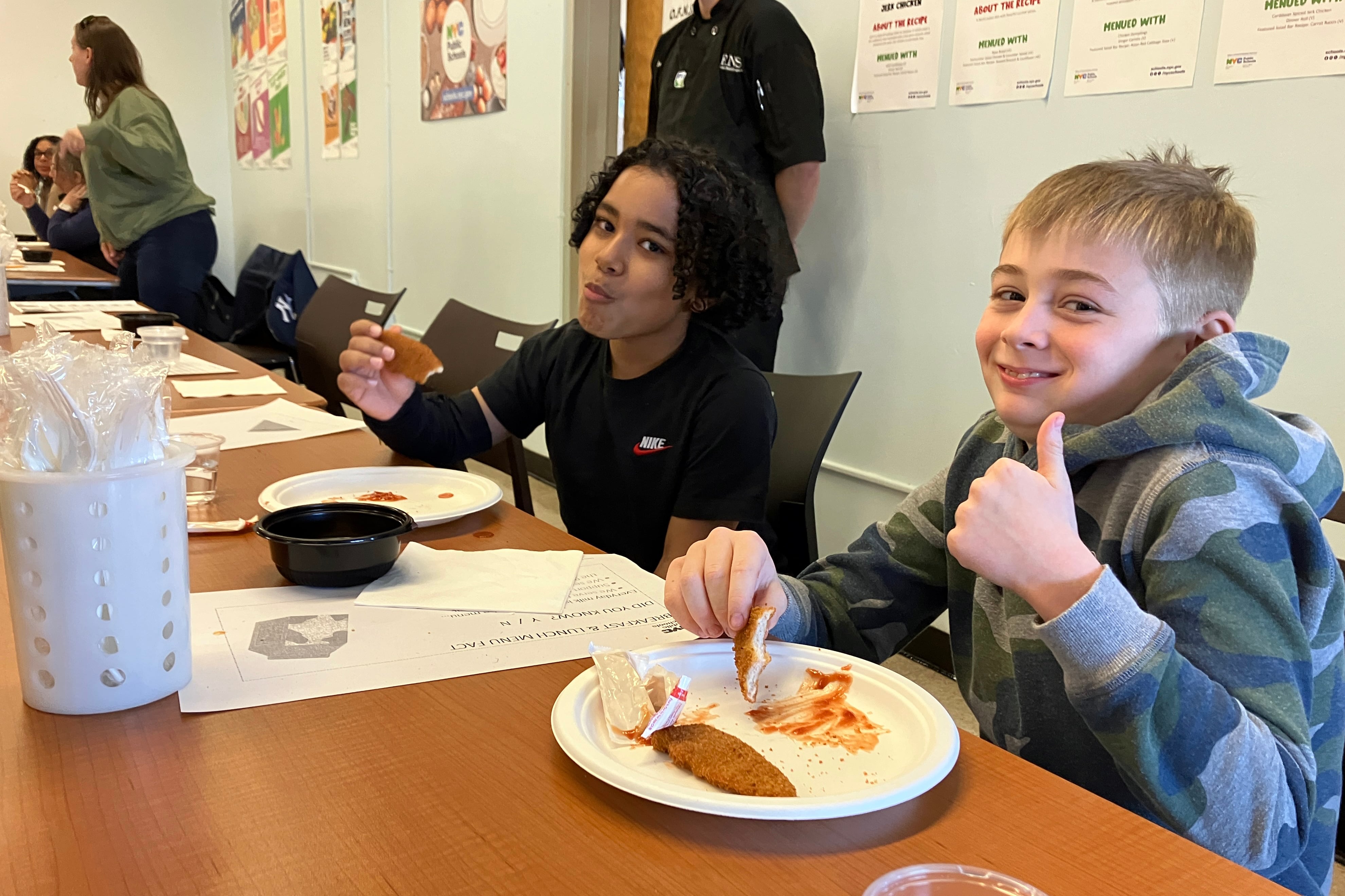 Two young students smile or give a thumbs up while eating from a plate of school lunch at a wooden table with two adults in the background.