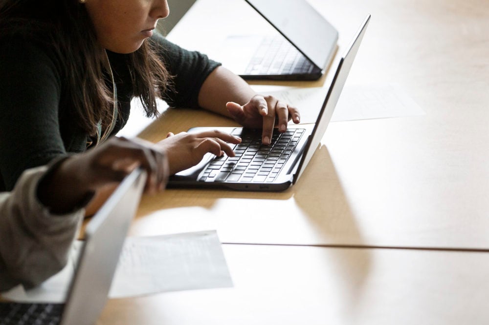 Two students sit at a wooden desk working on their laptops.