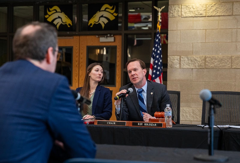 Two people sit at a black table with an American flag in the background and a view of a person's back in the foreground.