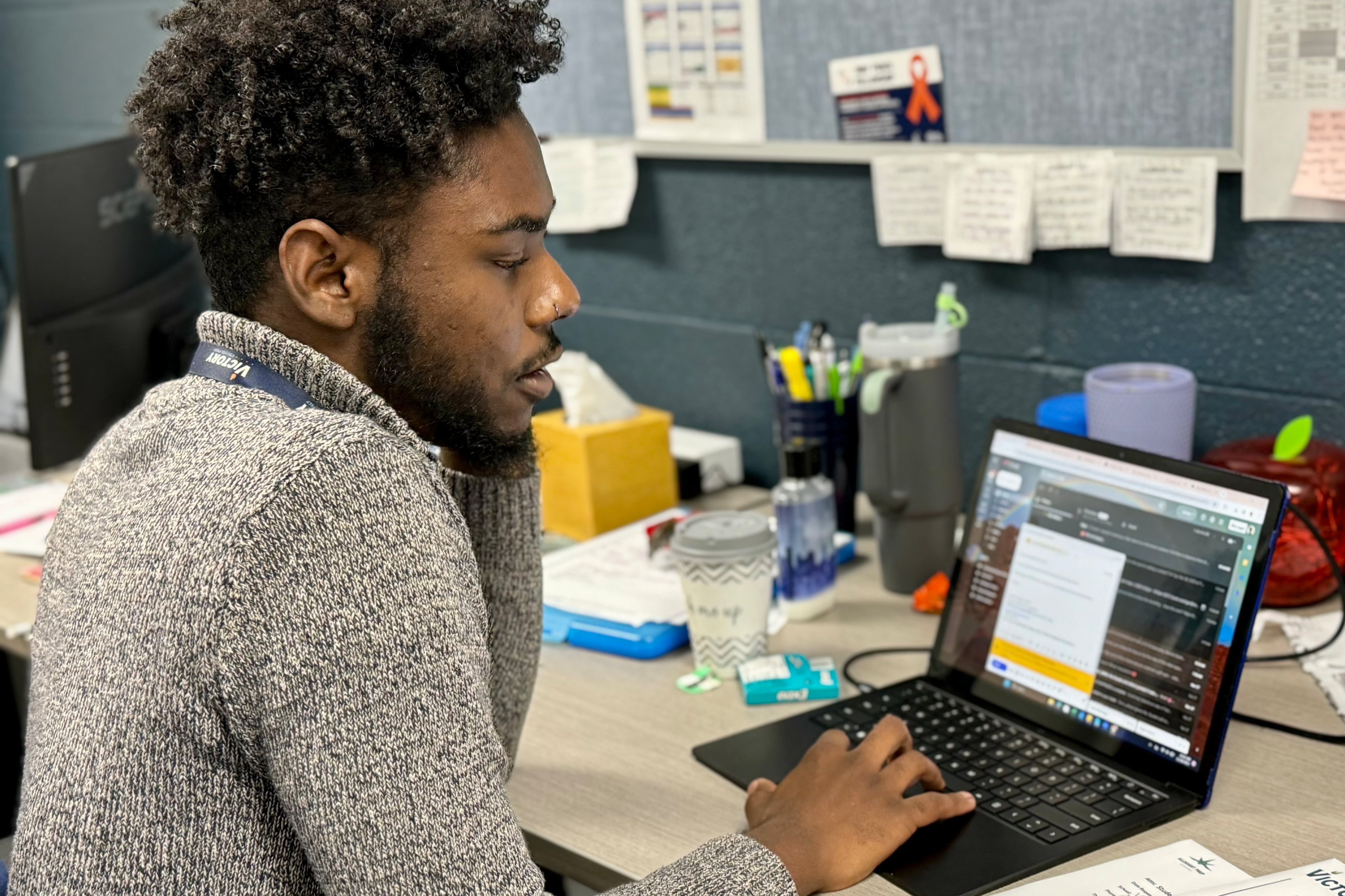 Man in gray sweater sits at desk and works on laptop computer.