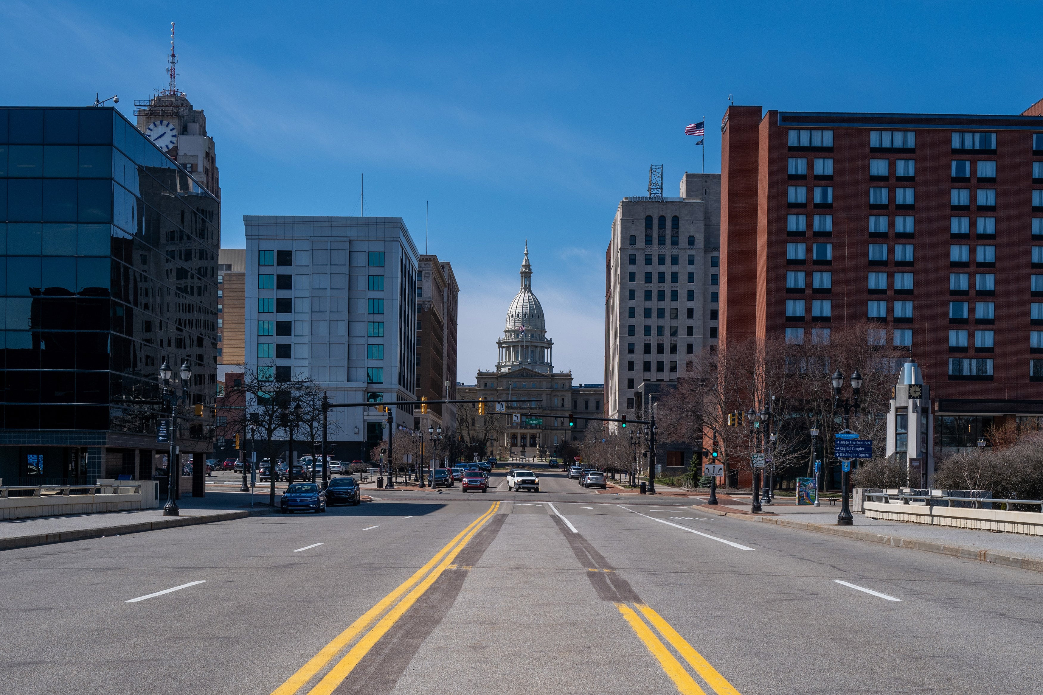 A view of Michigan's State Capitol building with a road in the foreground, buildings on both sides of the street and a blue sky in the background.