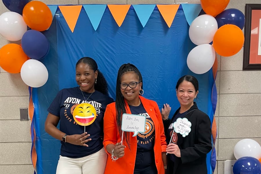 Three women pose in front of a blue backdrop with banners and balloons.