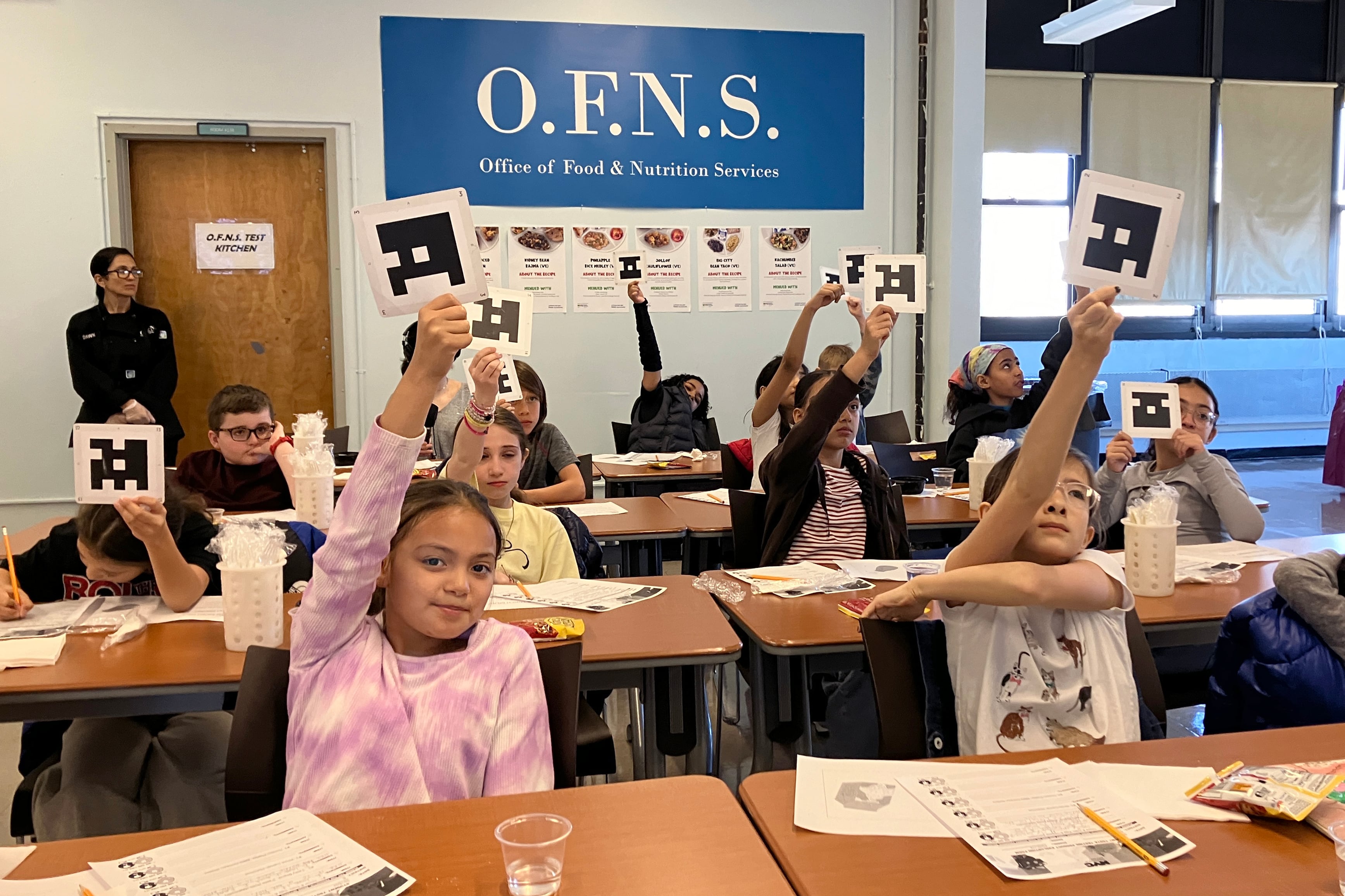 A group of young students sit at wooden desks with voting cards and sheets of paper in front of them.