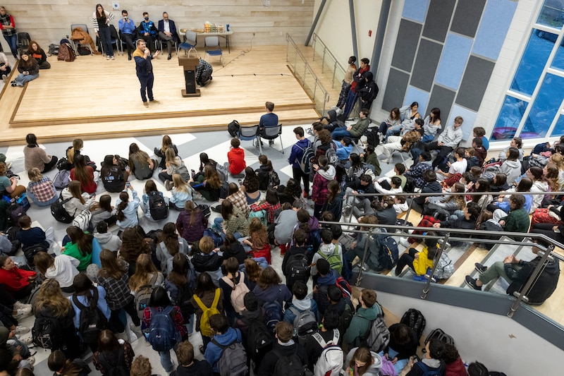 A person speaks on stage while a large crowd gathers around to listen.