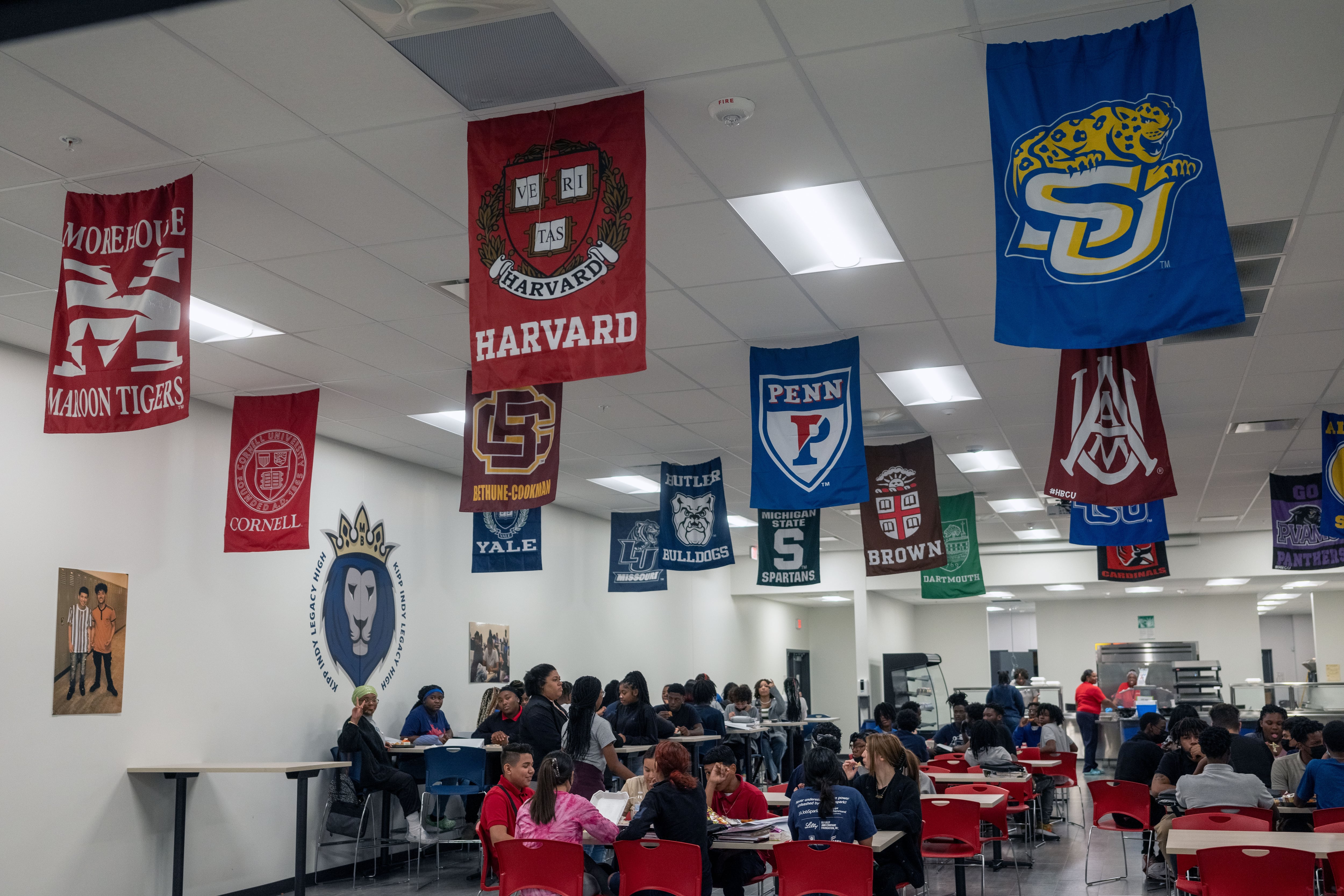 Flags of various colleges hang from the ceiling of a high school cafeteria as students sit at their tables.