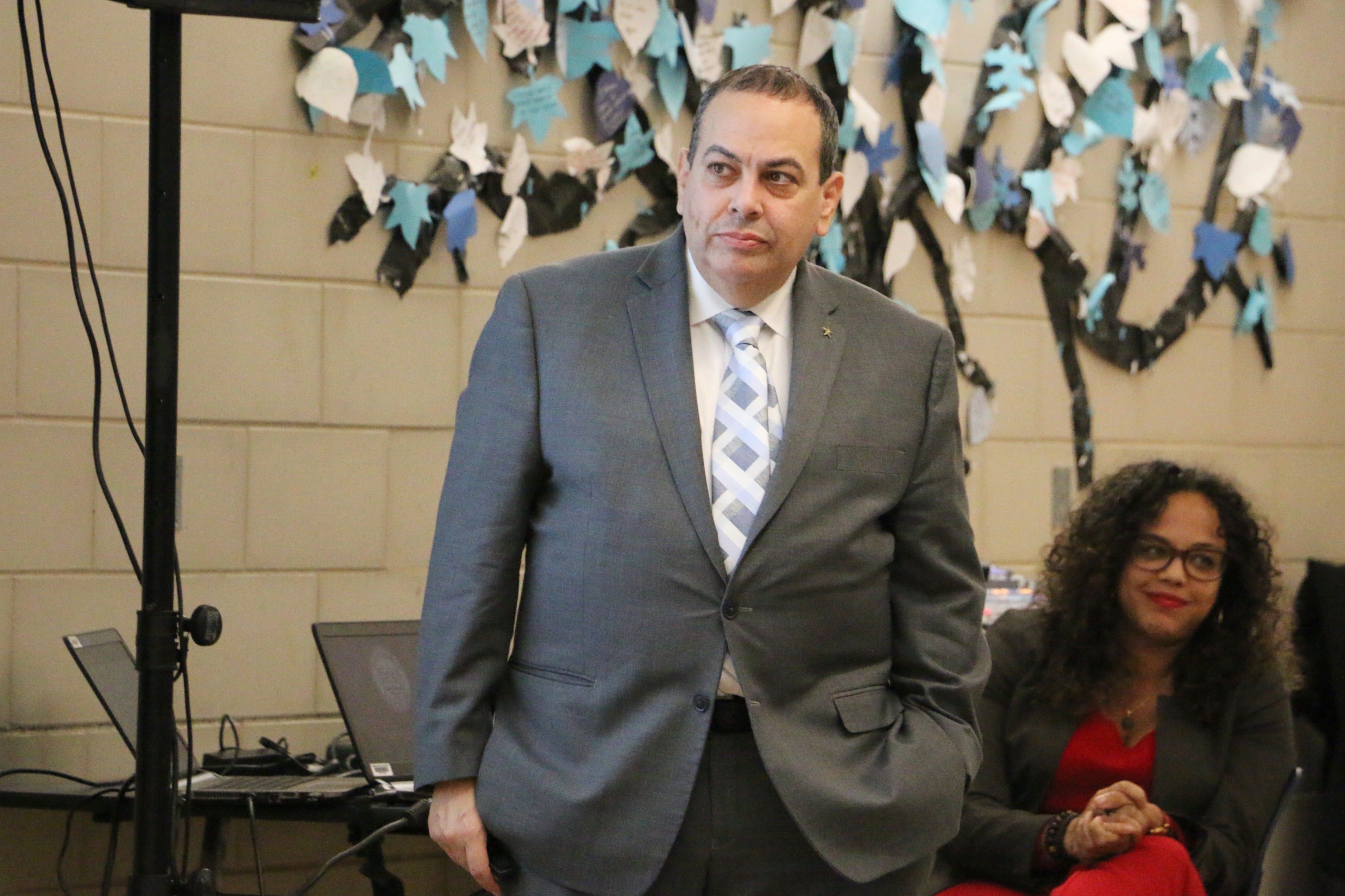 A man with a dark suit and a blue tie stands in front of another adult and a tan brick wall.