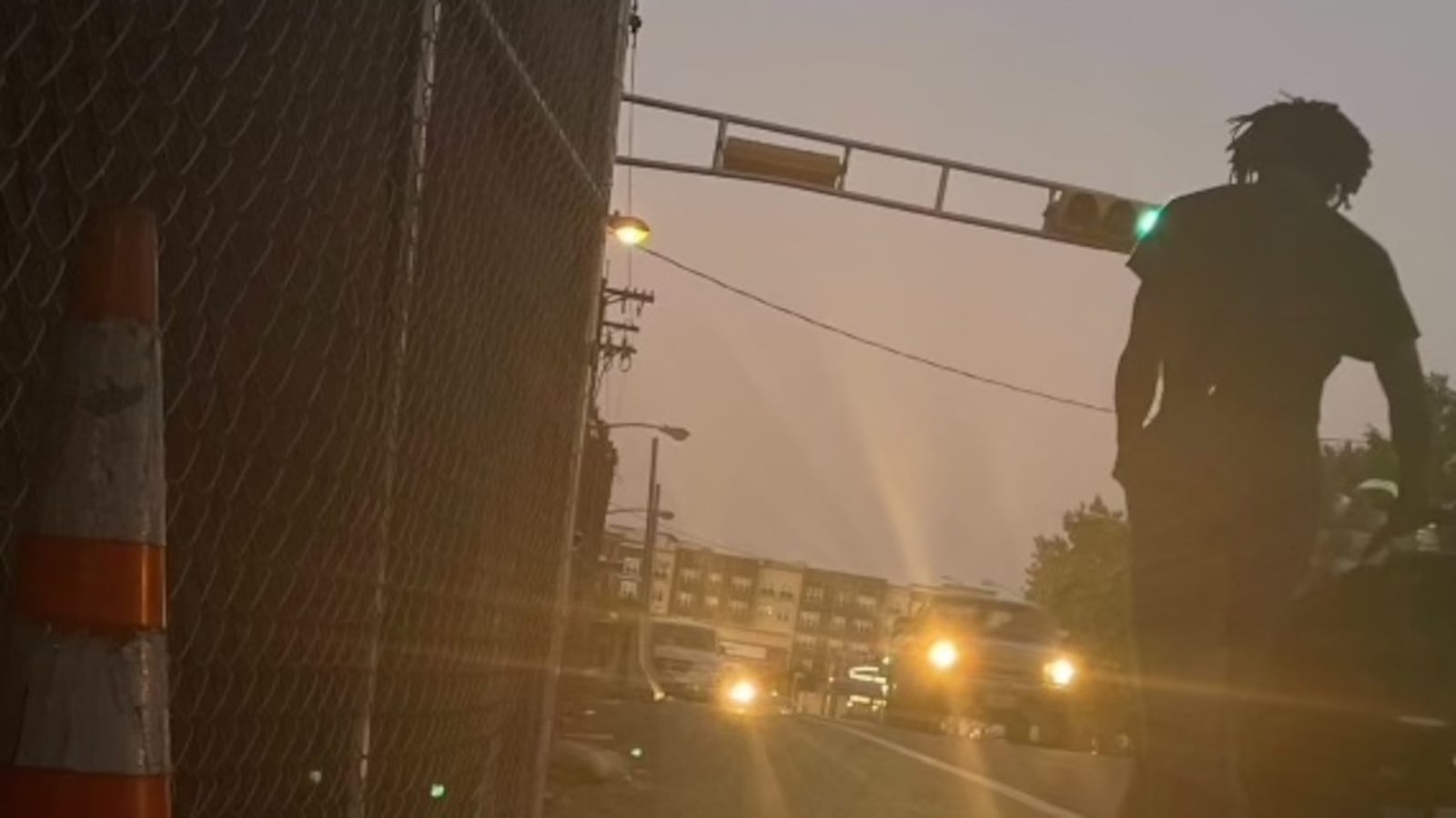 A streetscape at dusk. In the foreground is a person's silhouette. There are headlights in the background.