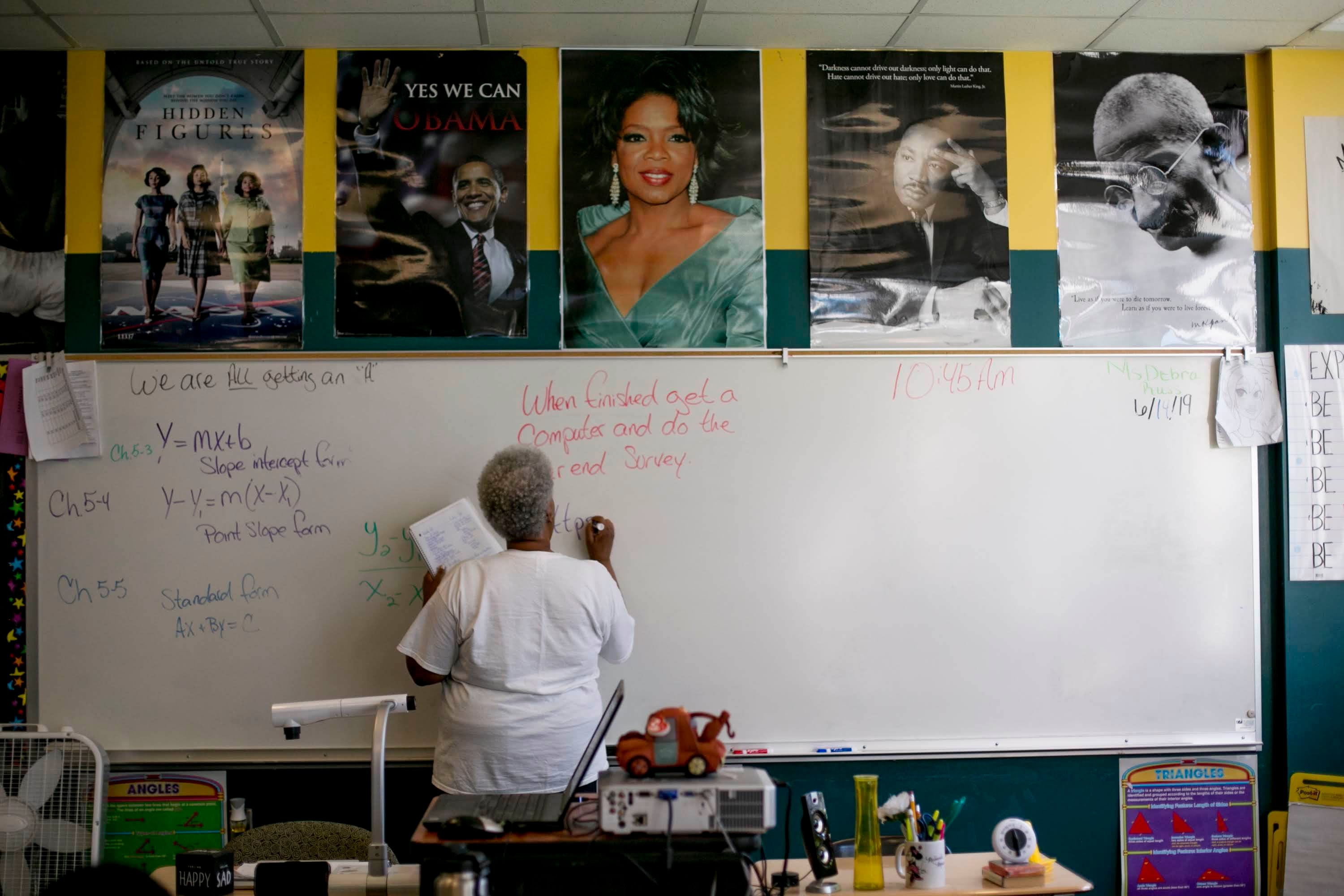 A teacher writes on a dry-erase board in a classroom. Math equations are written on the board to her left. Above the board are posters of Barack Obama, Oprah Winfrey, Martin Luther King Jr., Mahatma Gandhi, and the cast of the movie Hidden Figures.