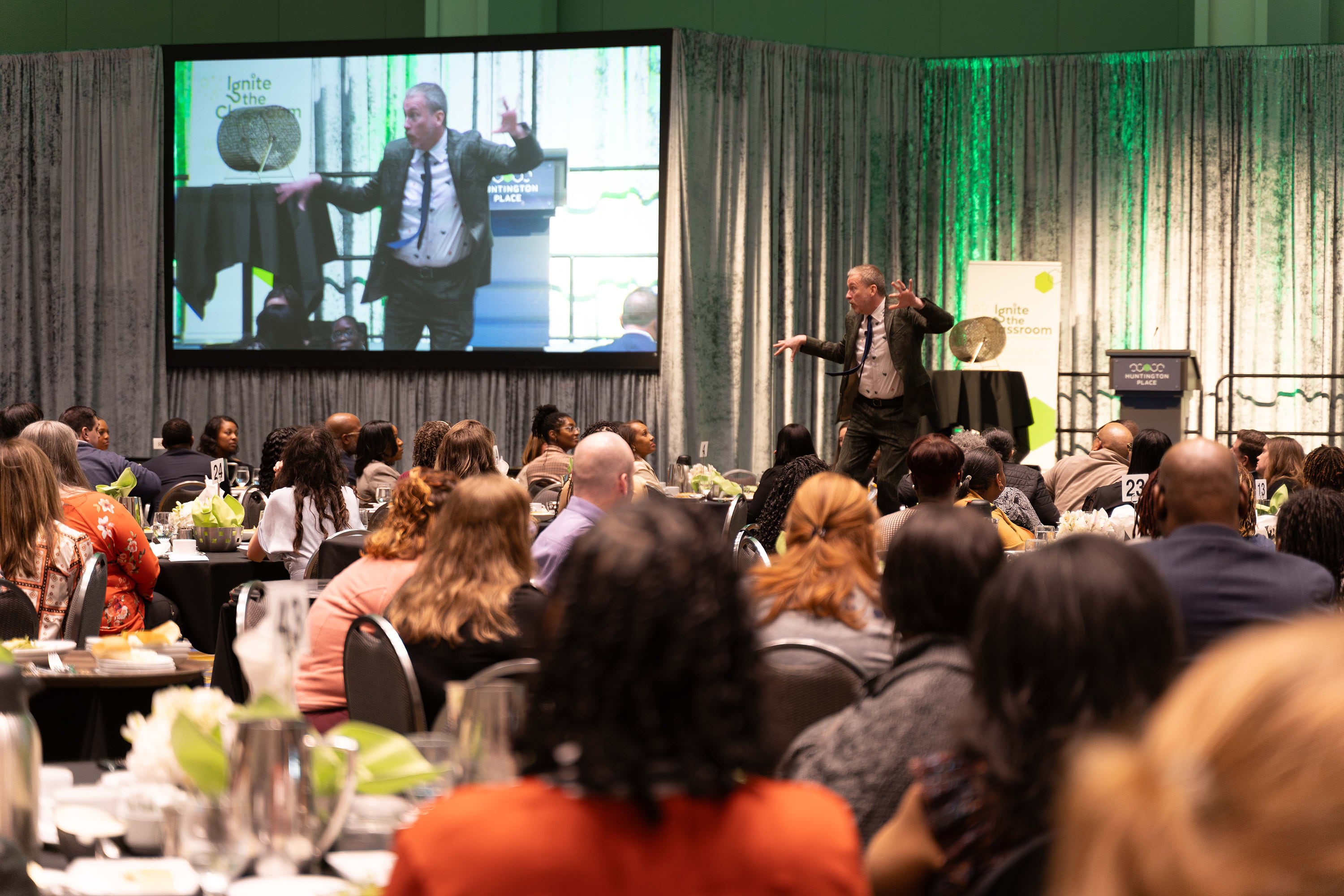 An adult stands on stage while a room full of adults sit at round tables in a banquet hall.