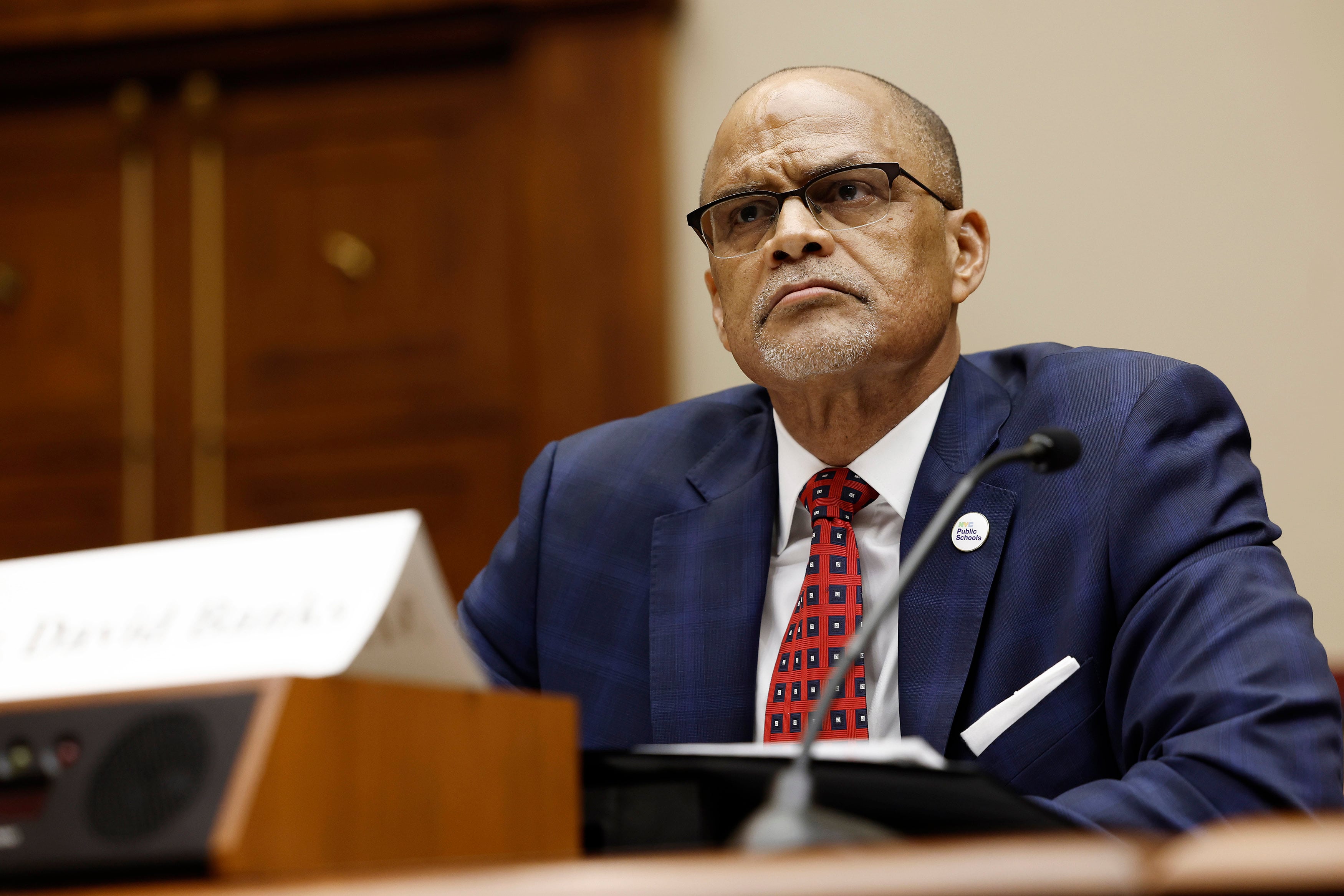 A man with no hair and wearing glasses and a dark blue suit with a red tie sits at a wooden desk staring ahead with a name plate and microphone in the foreground.