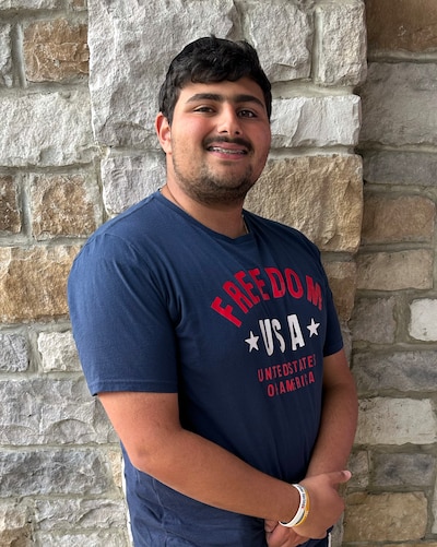 A high school student with short dark hair and wearing a blue t-shirt with red and white writing poses for a photograph in front of a stone wall.