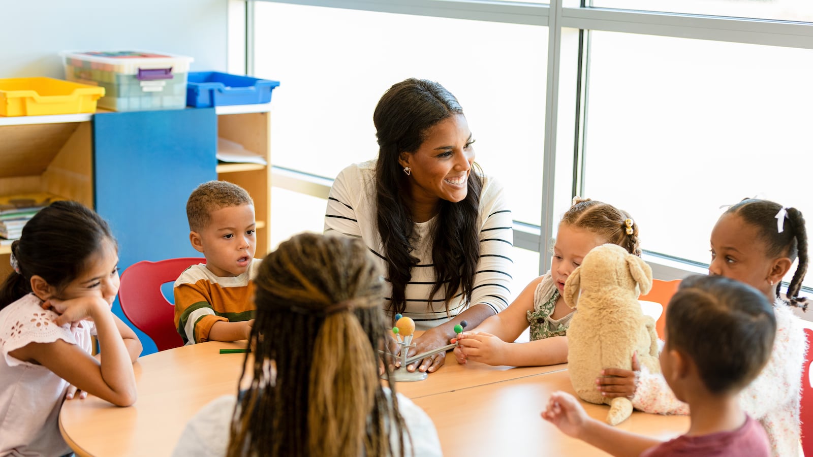 A group of young children sit around a round table as their teacher teaches them using a model of the solar system.