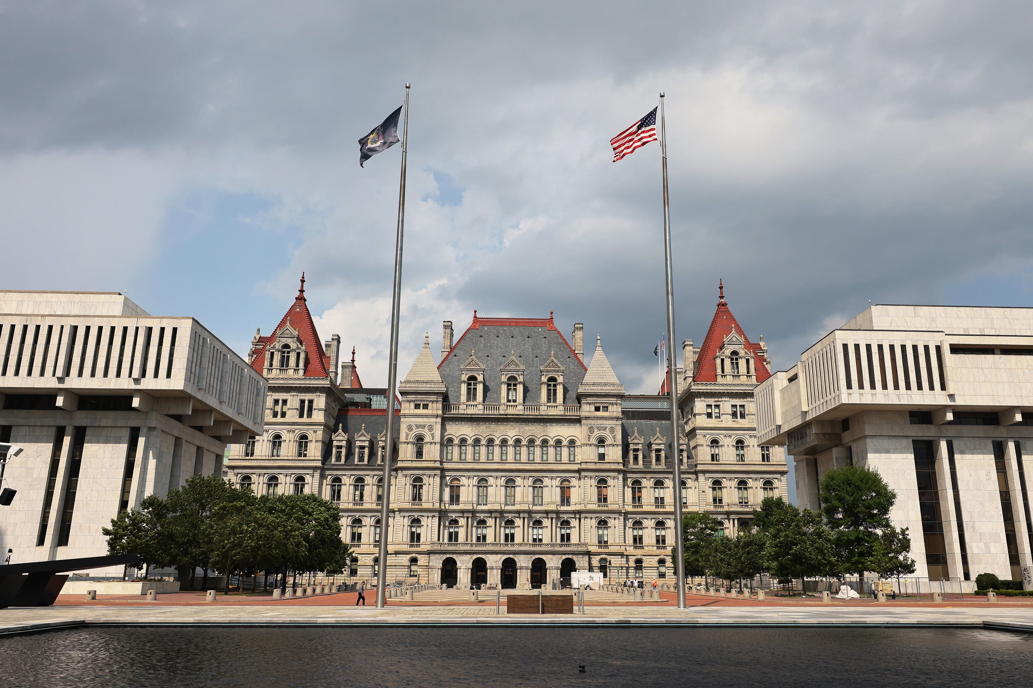 Two flags fly above a large tan stone building with a reflection pond in the foreground and a cloudy sky in the background.