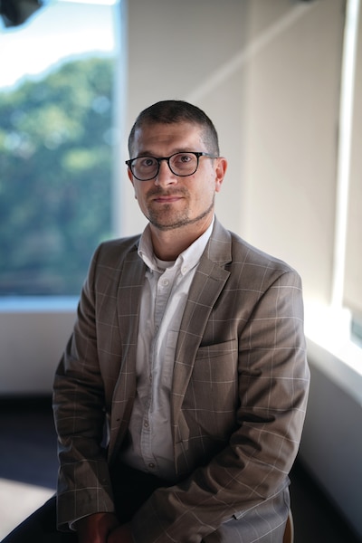 A person wearing glasses and a tan suit sits and looks directly at the camera to pose for a portrait.