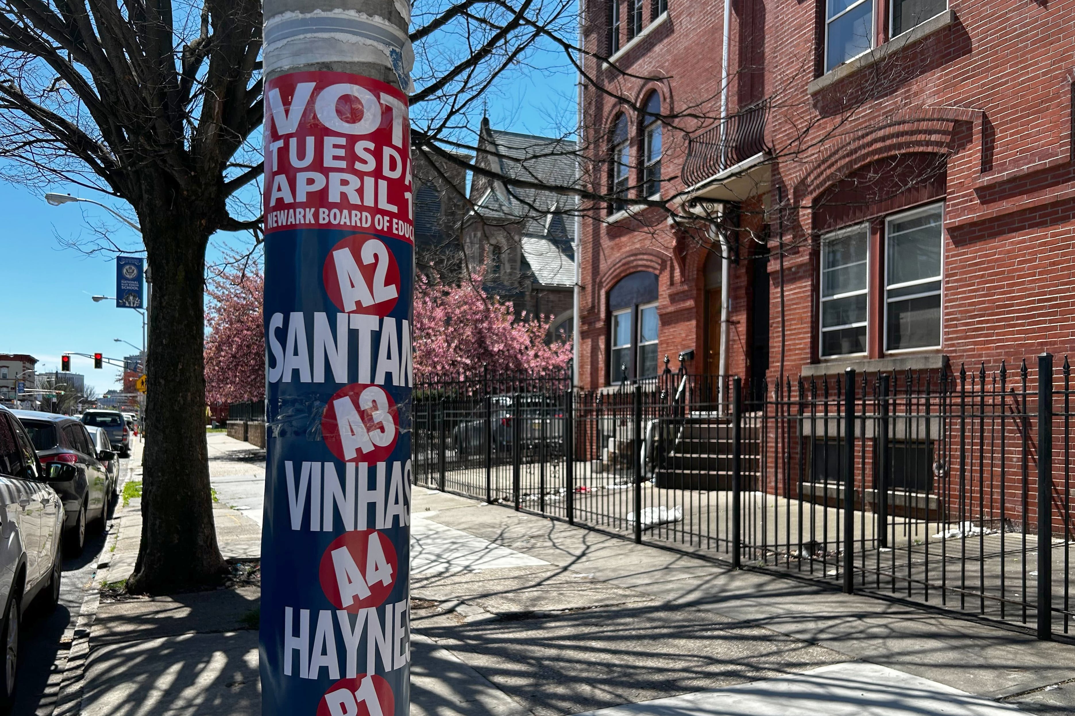 A street pole covered in candidate signs on a sidewalk in front of a red brick building.