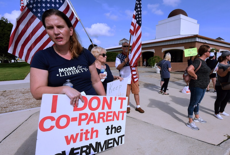 People protest outside holding signs with American flags and a building in the background.