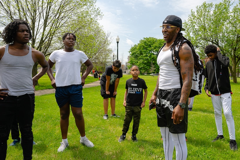 An adult wearing a ball cap and a white tank top stands to the right of a group of students all in work out clothes outside.