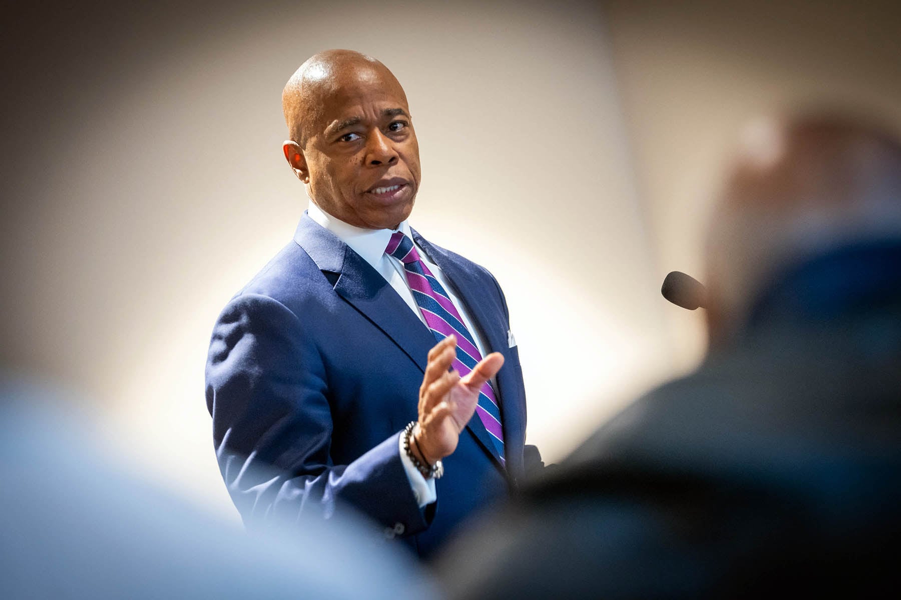 A man with no hair and wearing a dark suit, speaks at a podium with his hand up. A white wall in the background.