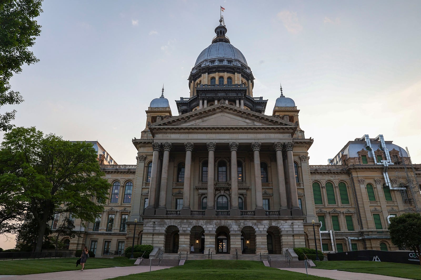 A large stone state capitol building with trees and grass on both sides and in the foreground. A light blue sky with a few clouds in the background.