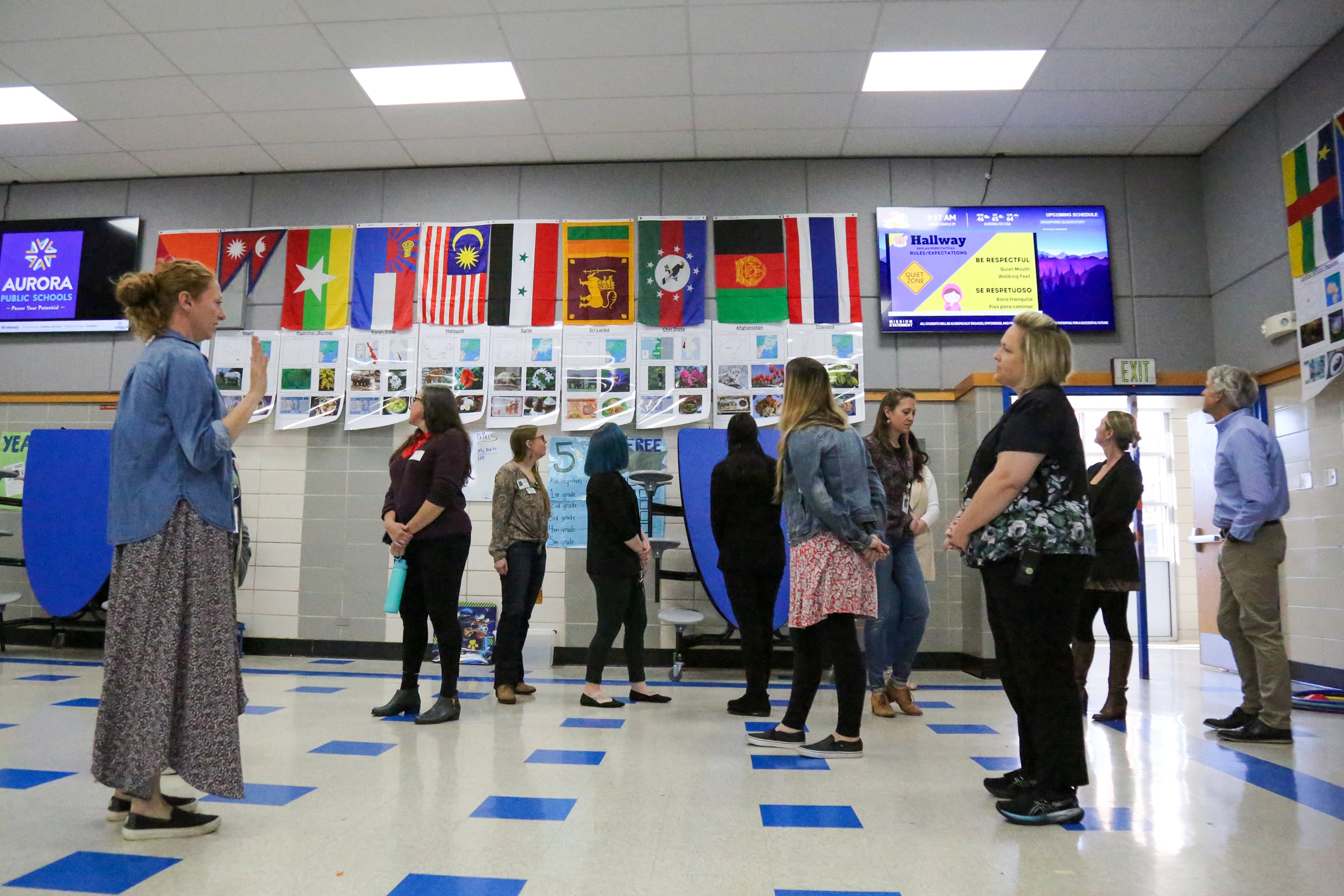 A group of people stand in a large room with flags from different places lining the wall in the background.