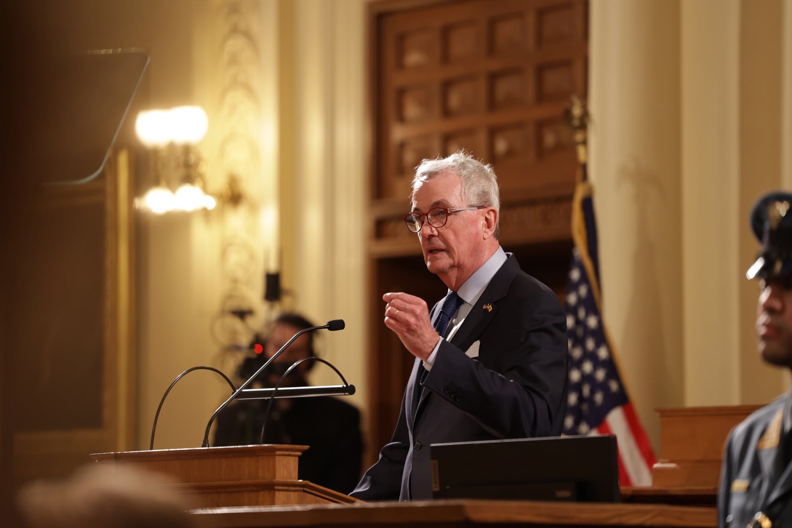 A man with short white hair stands at a wooden podium with an American flag on the side and a wooden door in the background.