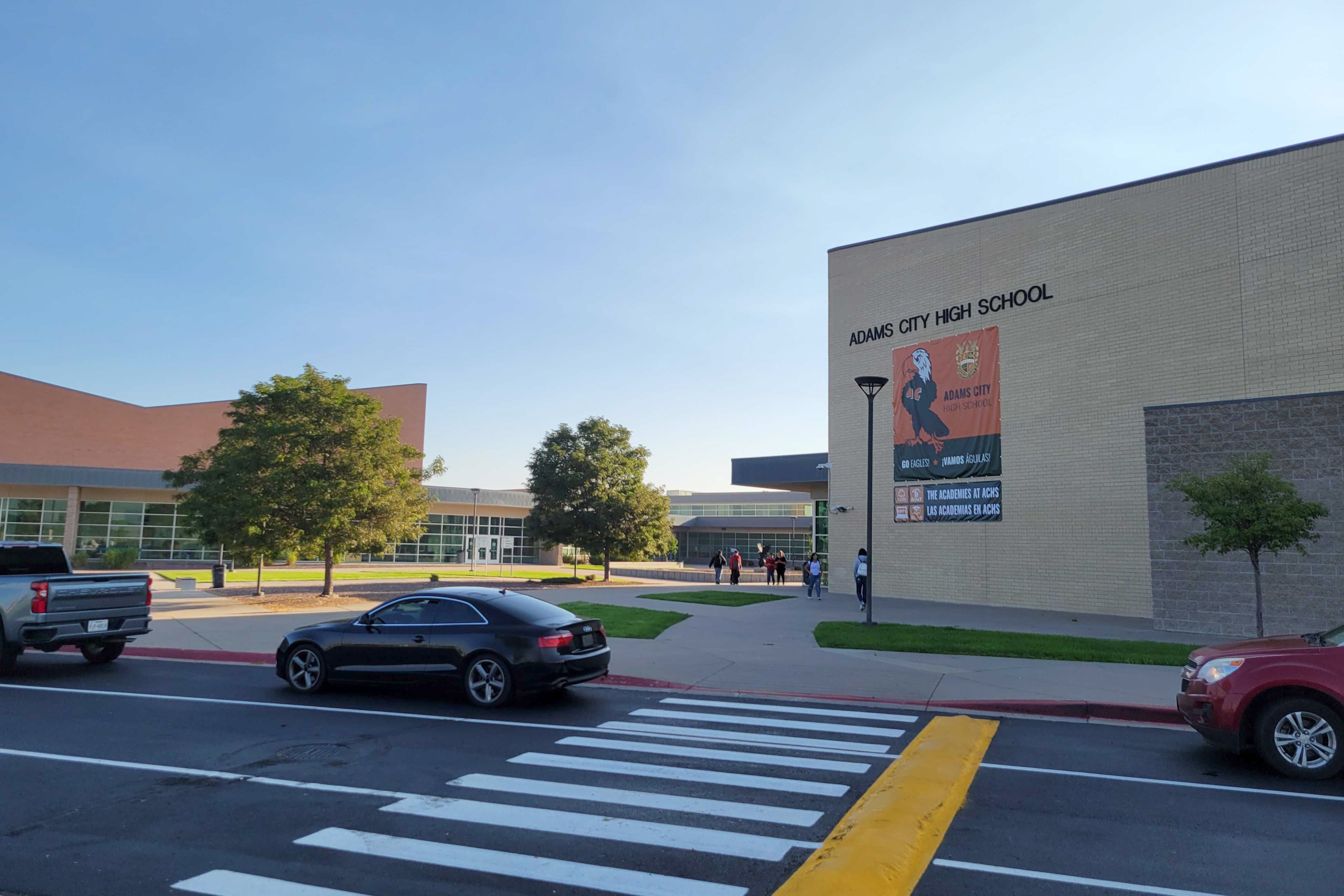 Two large brick school buildings with students walking along the sidewalk between them and cars parked on the street in the foreground.