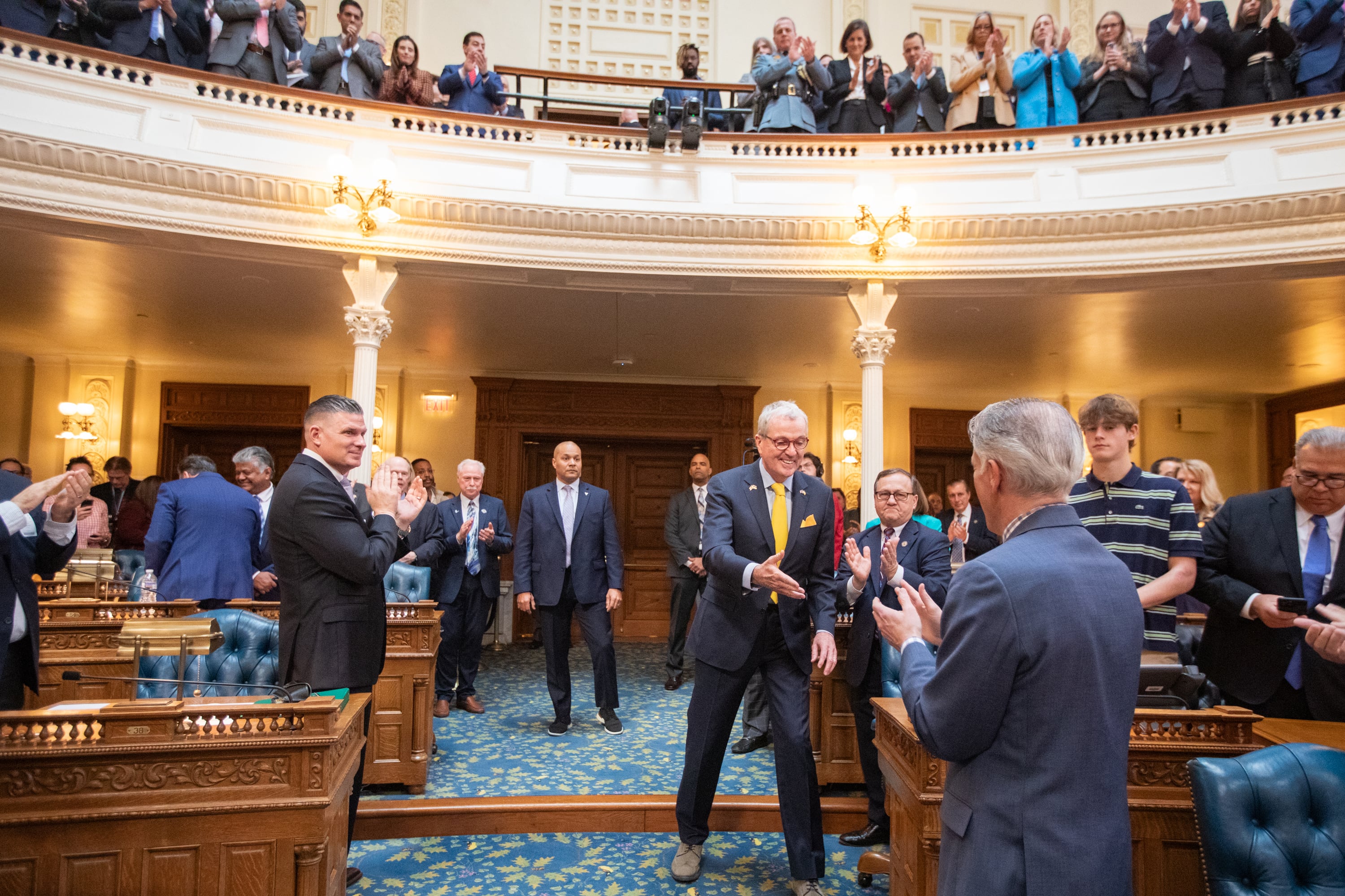 A man wearing a dark suit goes to shake someone else's hand with a large crod in the background.