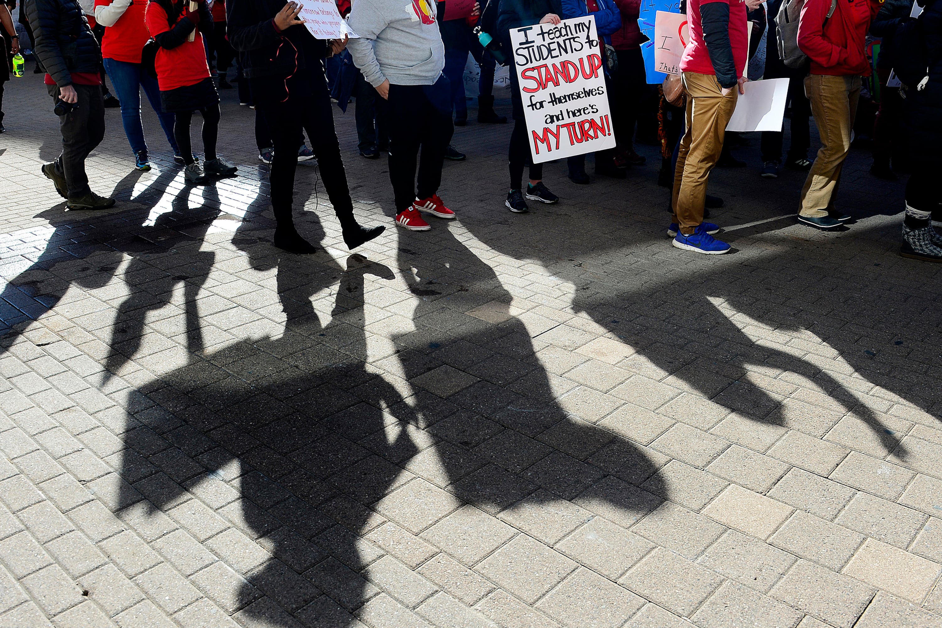 Shadows of people walking during a protest and holding up signs outside on tan brick sidewalk.