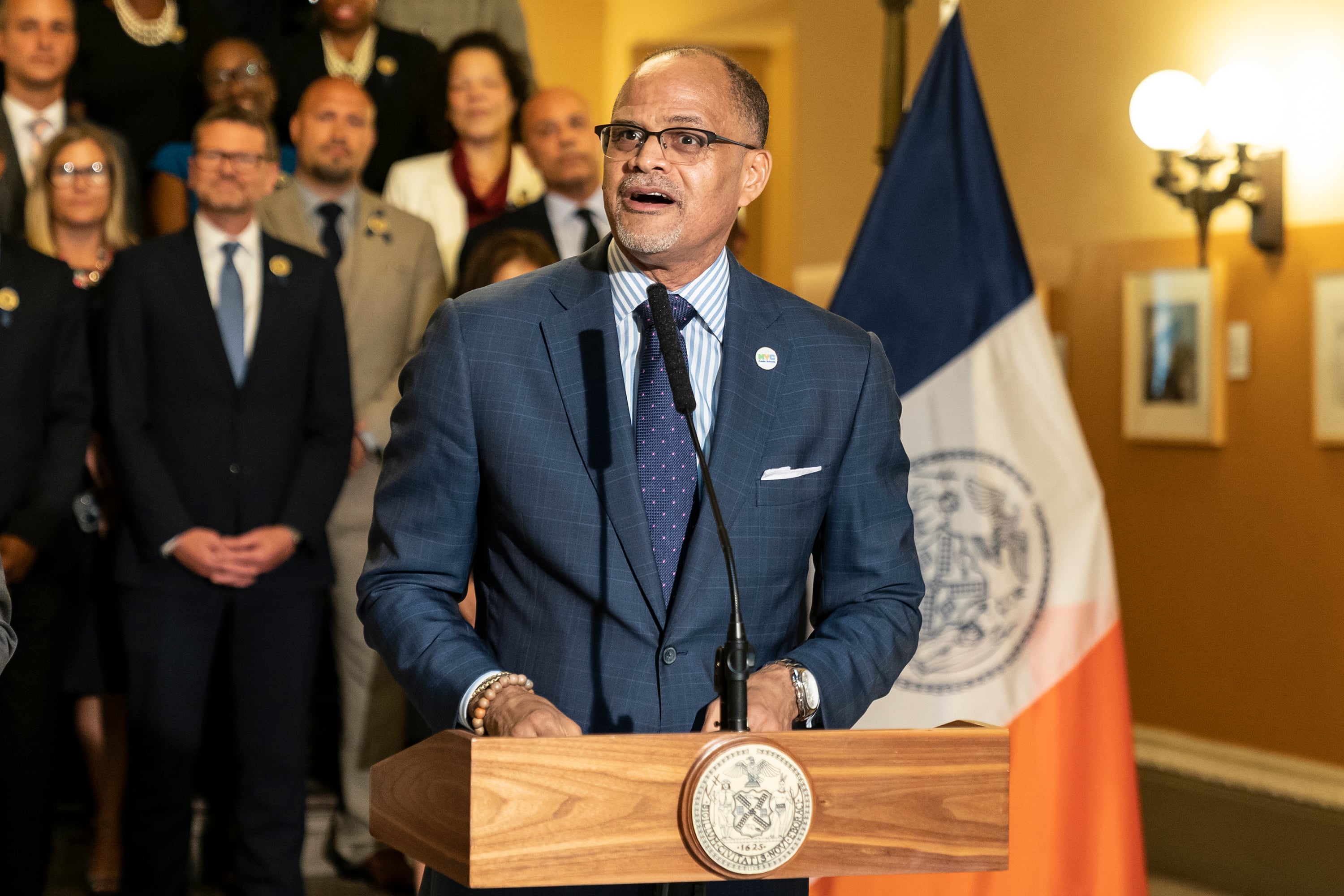 A man wearing a suit and glasses speaks from a wooden podium with a group of people standing behind him.