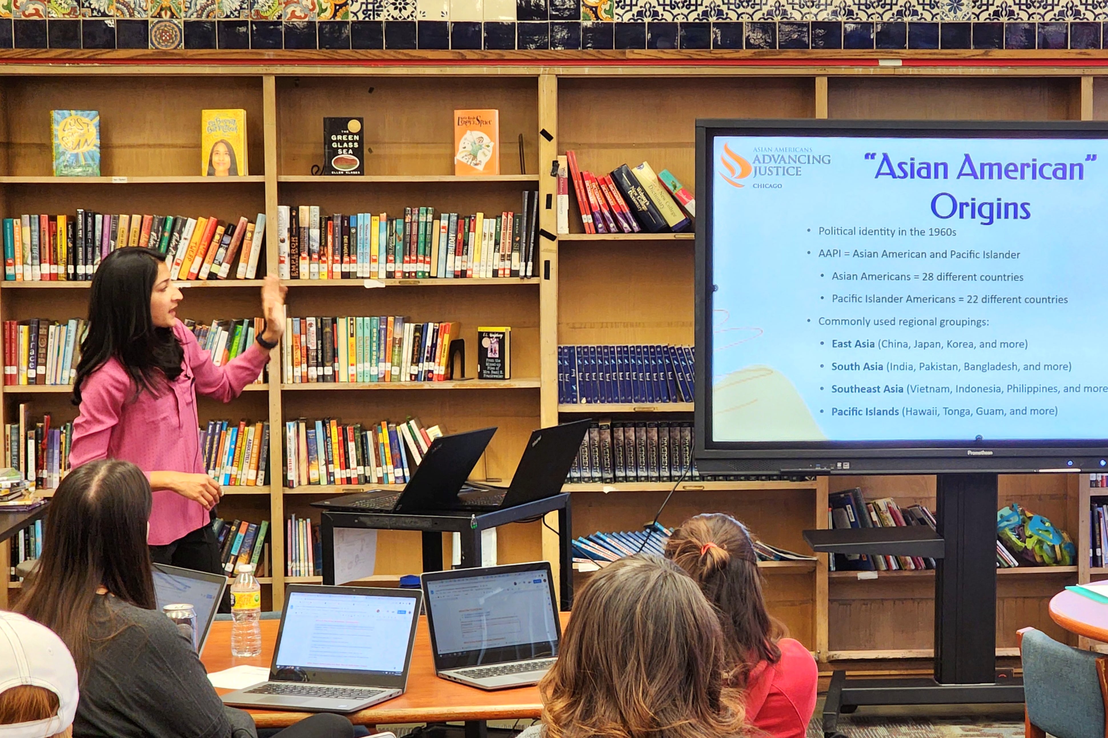 A woman in pink, at left, speaks with text projected on a screen in front of shelves of books.