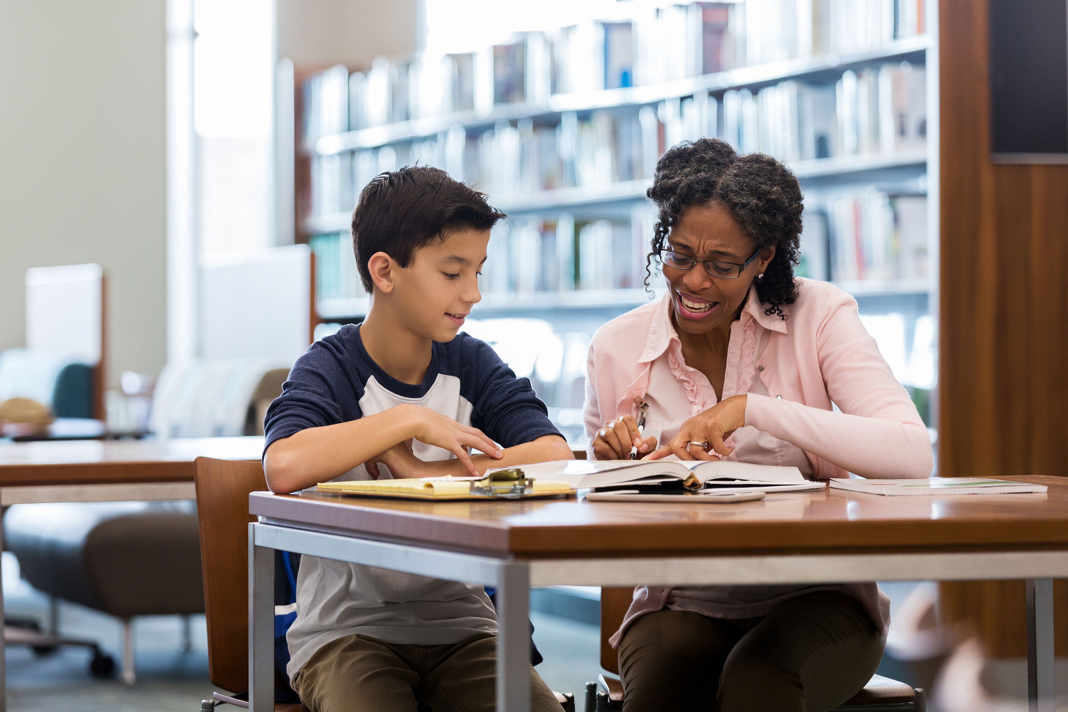 A middle school student sits next to an adult librarian at a wooden desk reading from a book with a shelf full of books in the background.